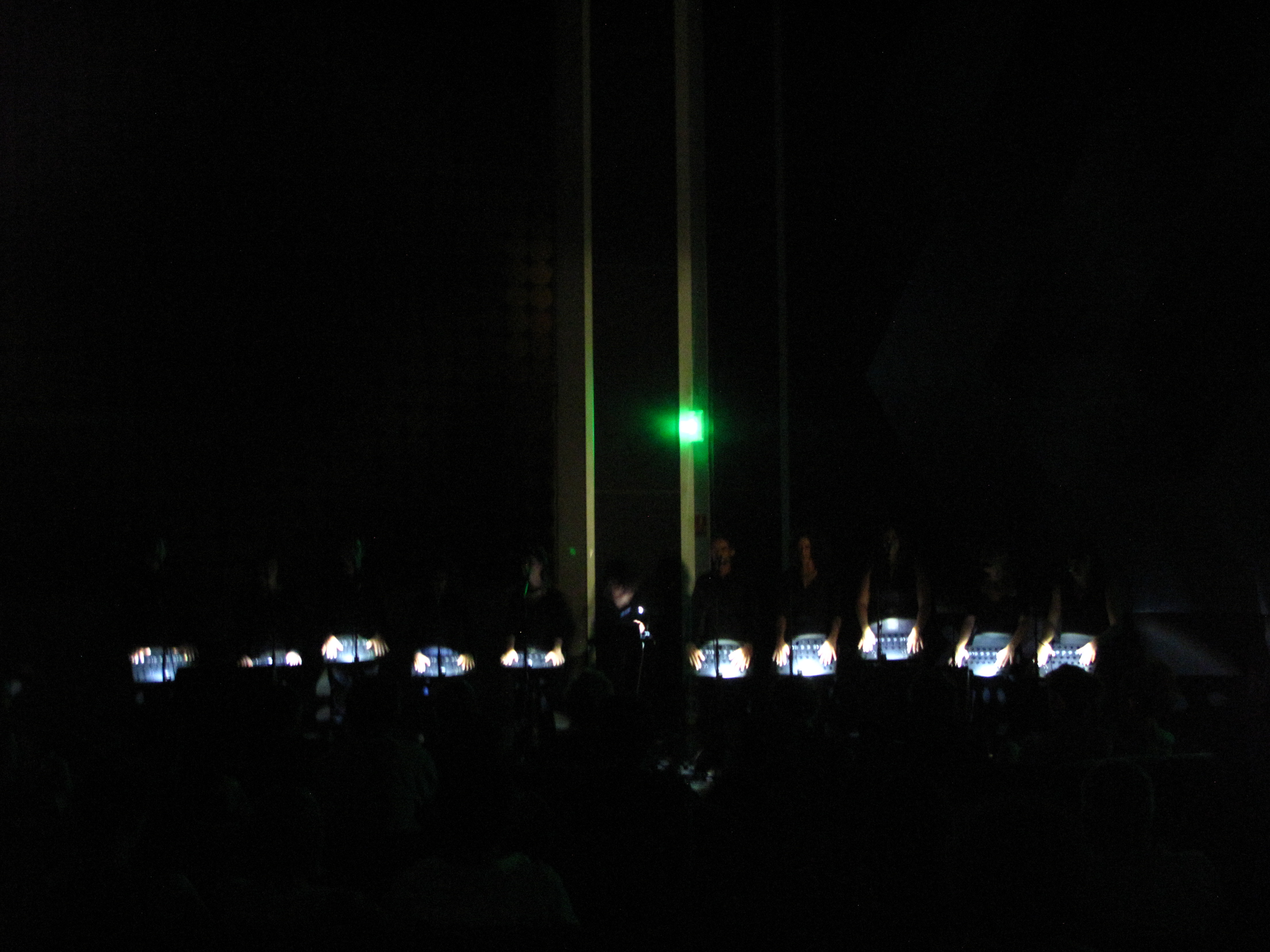 Choir on a dimly lit stage with illuminated music stands and audience silhouettes in the foreground.