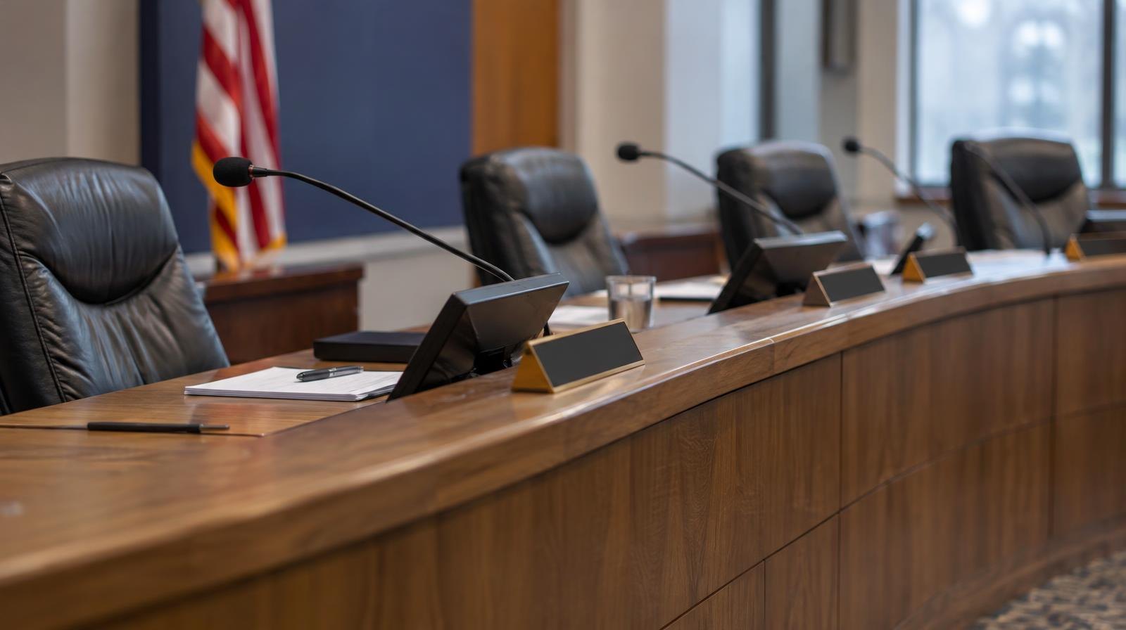 Empty school board meeting room with a curved dais, microphones, and chairs.