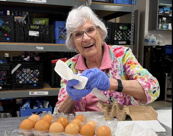 Elderly woman in floral jacket and blue gloves smiles while sorting eggs at a food pantry.