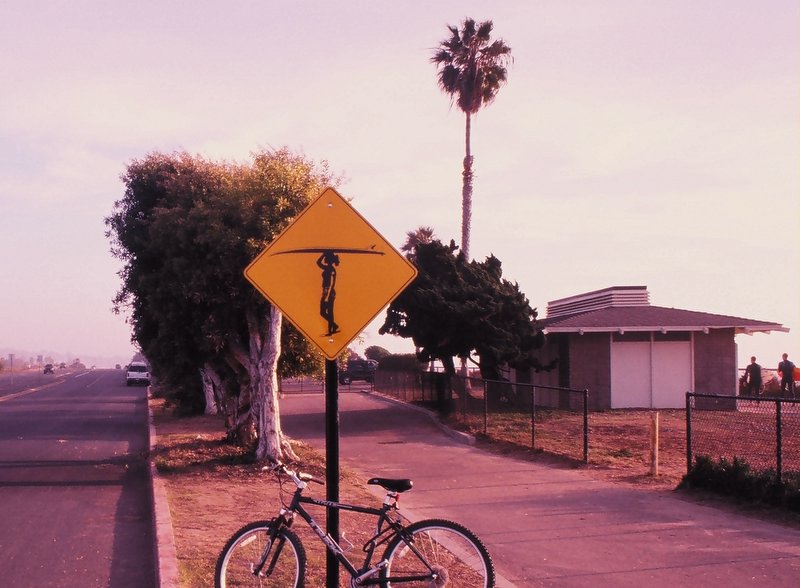Yellow surfer-crossing sign with a bicycle leaned against the post on a roadside.