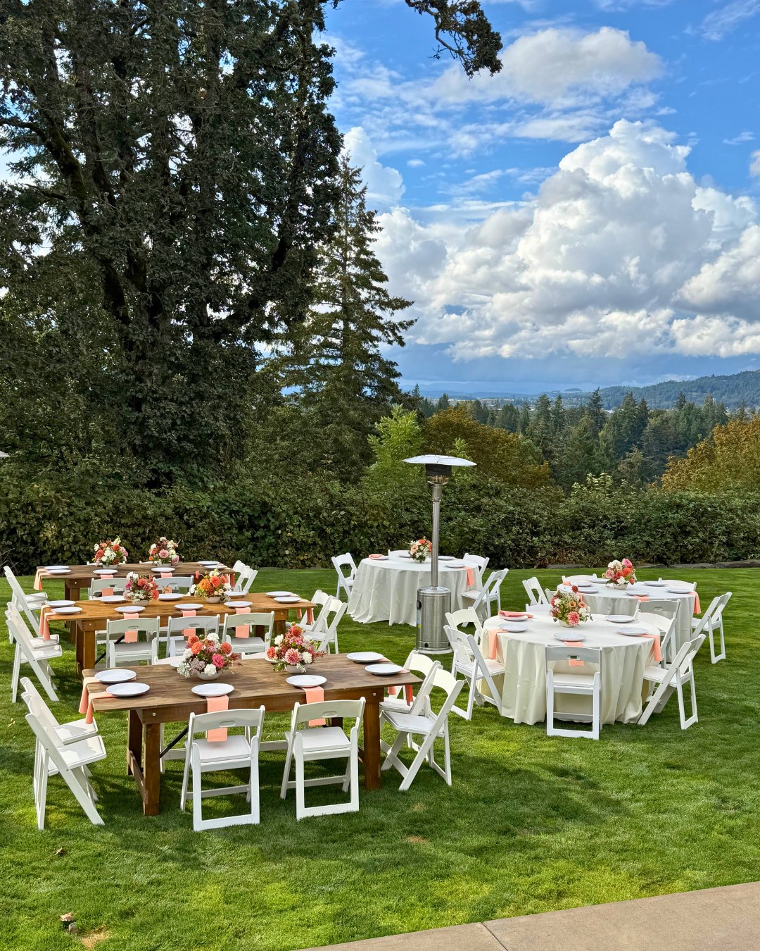 Outdoor event tables with white linens and floral centerpieces on a grass lawn beneath a partly cloudy sky.