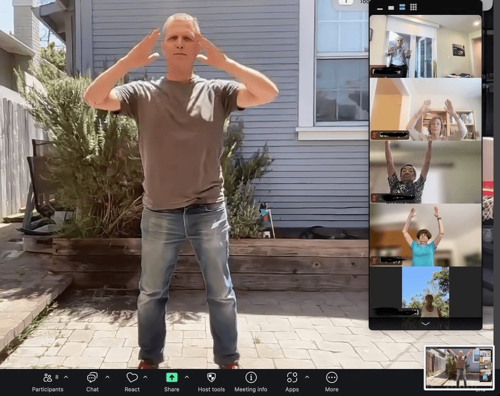 A man outdoors raises his hands to his head while a tablet shows Zoom participants doing the same.