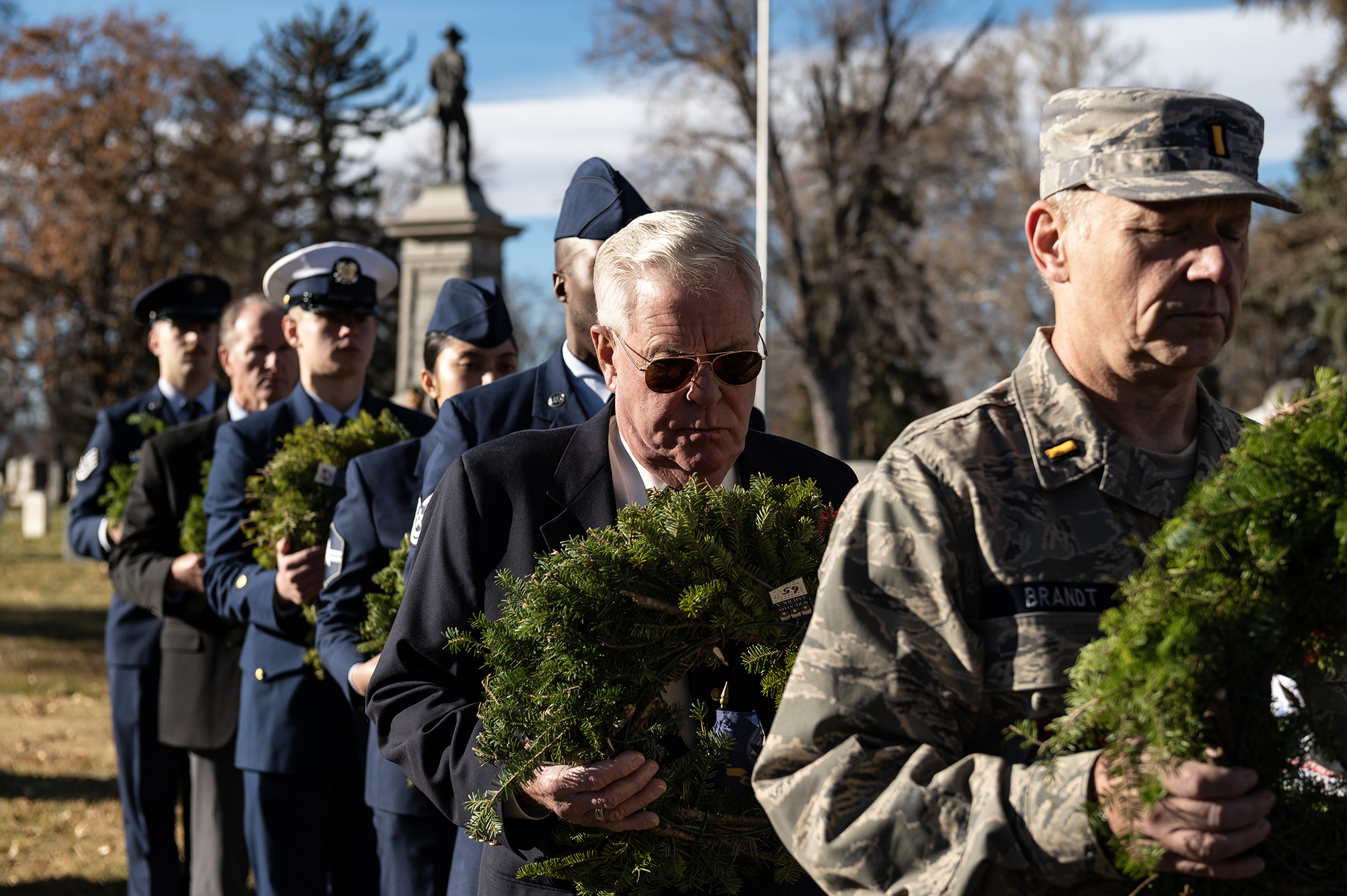 Military volunteers in uniform carry evergreen wreaths through a cemetery.