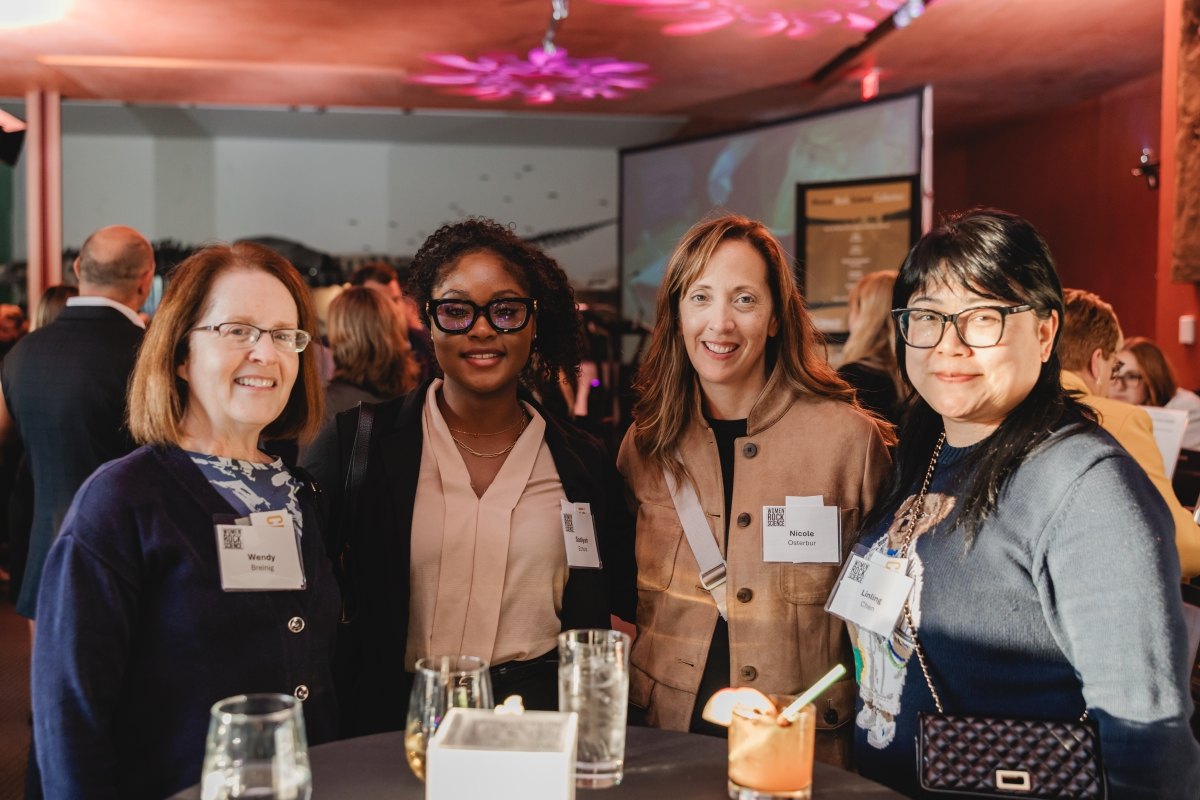 Four women smiling together at an indoor event, wearing name badges, with drinks on a table.
