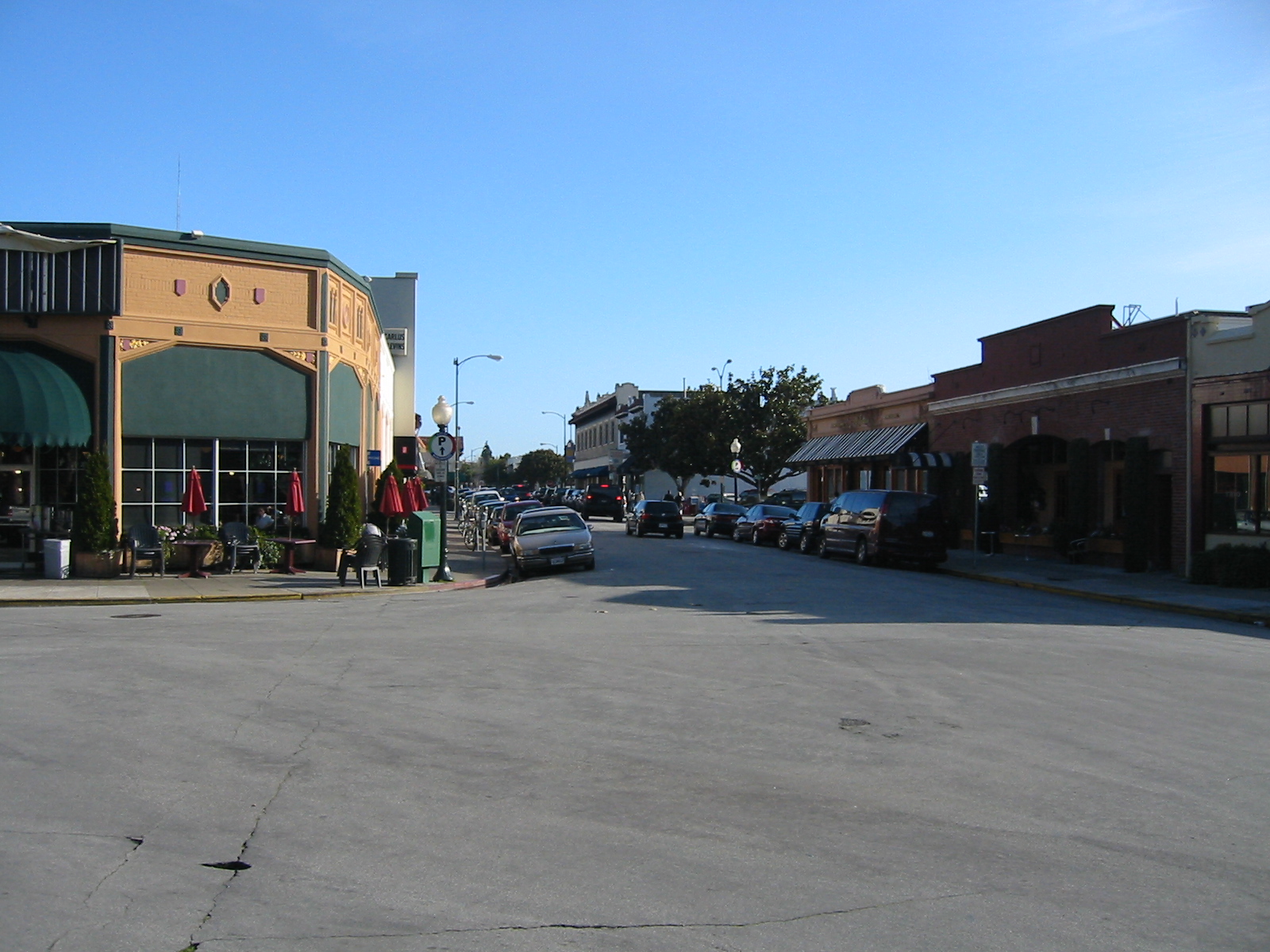 Downtown street lined with commercial storefronts, parked cars, and street lamps under a blue sky.