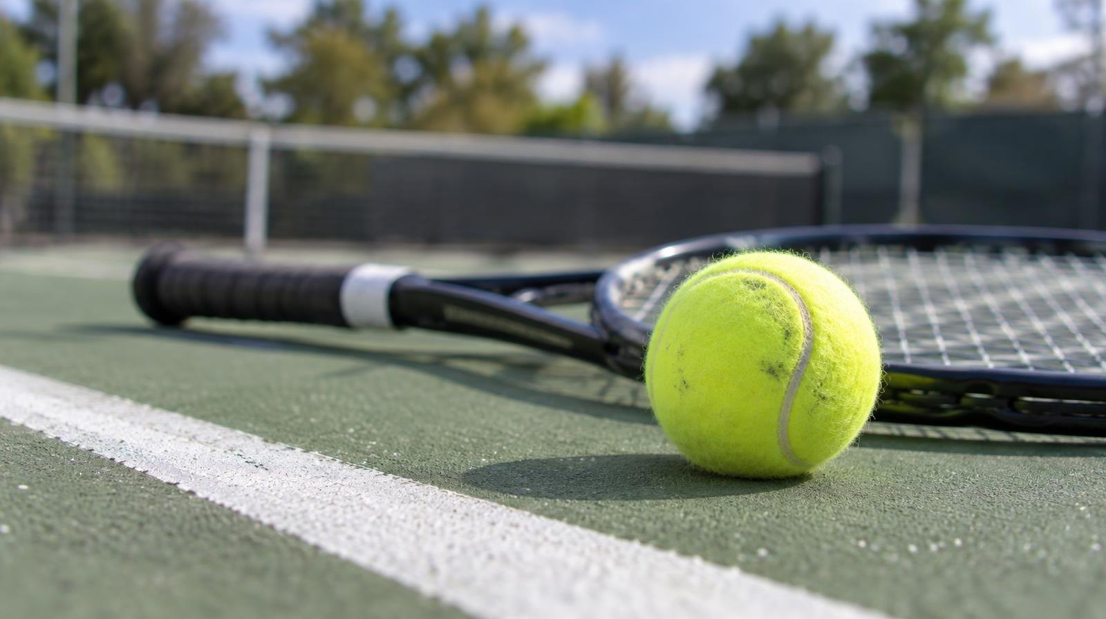 Yellow tennis ball resting on a green court beside a tennis racket.