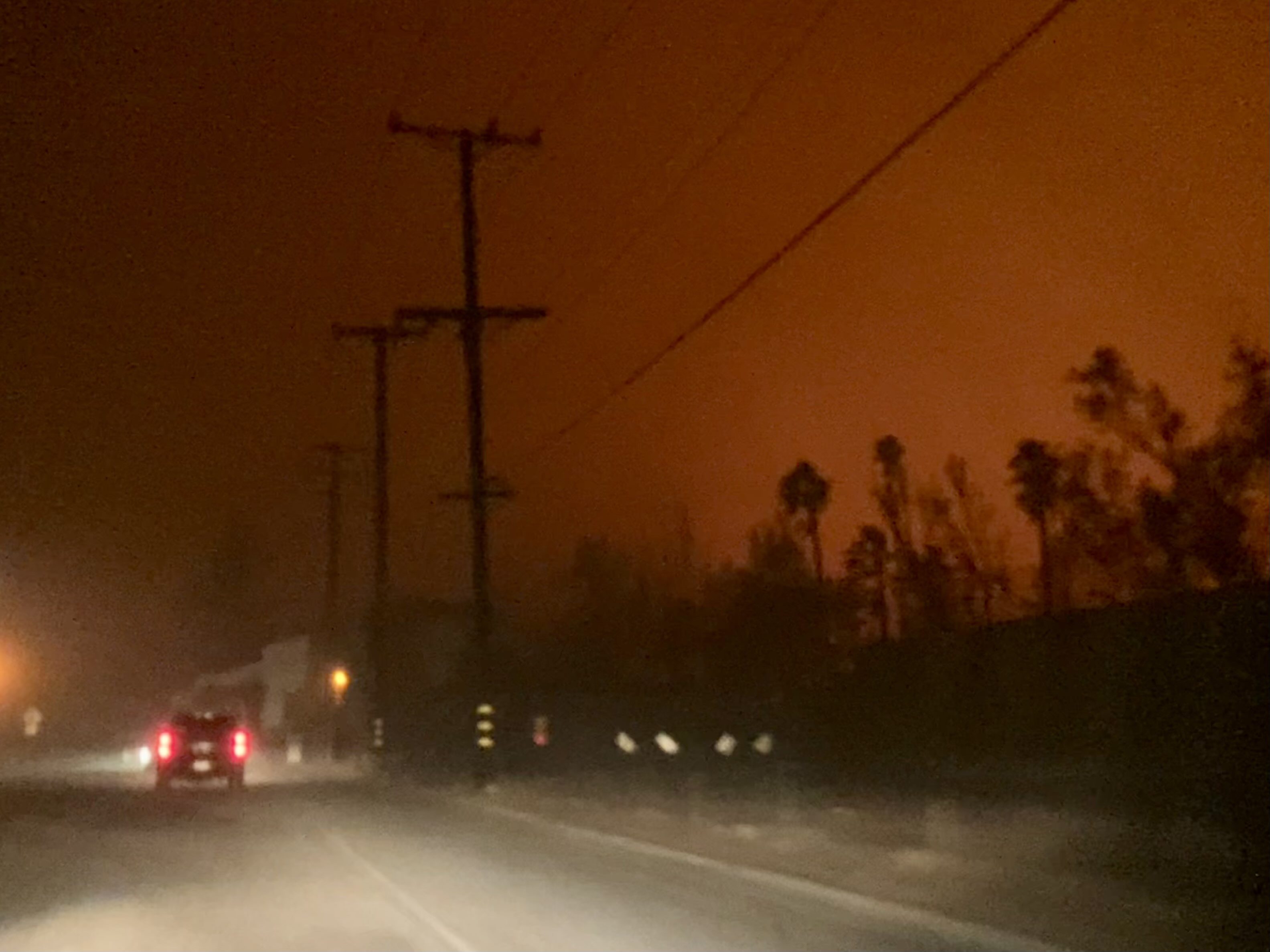 Orange-red smoke fills the night sky above a Pasadena street with utility poles and palm trees silhouetted against the fire's glow.