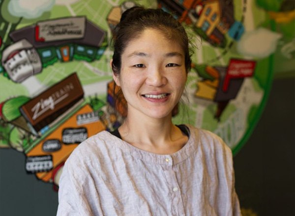 A smiling woman stands in front of a colorful illustrated map of Zingerman's businesses.