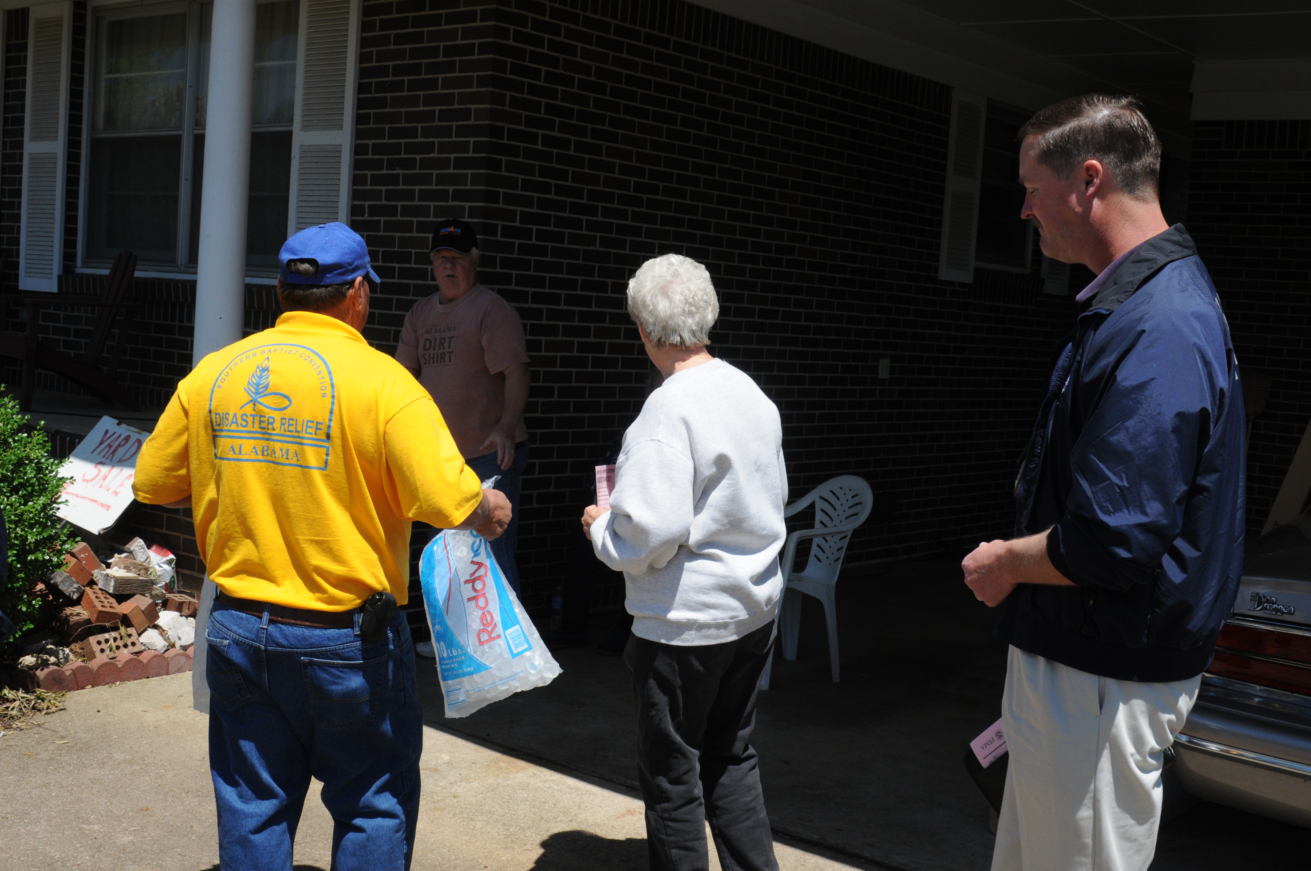 Three people stand outside a brick building, one holding a bag of ice.