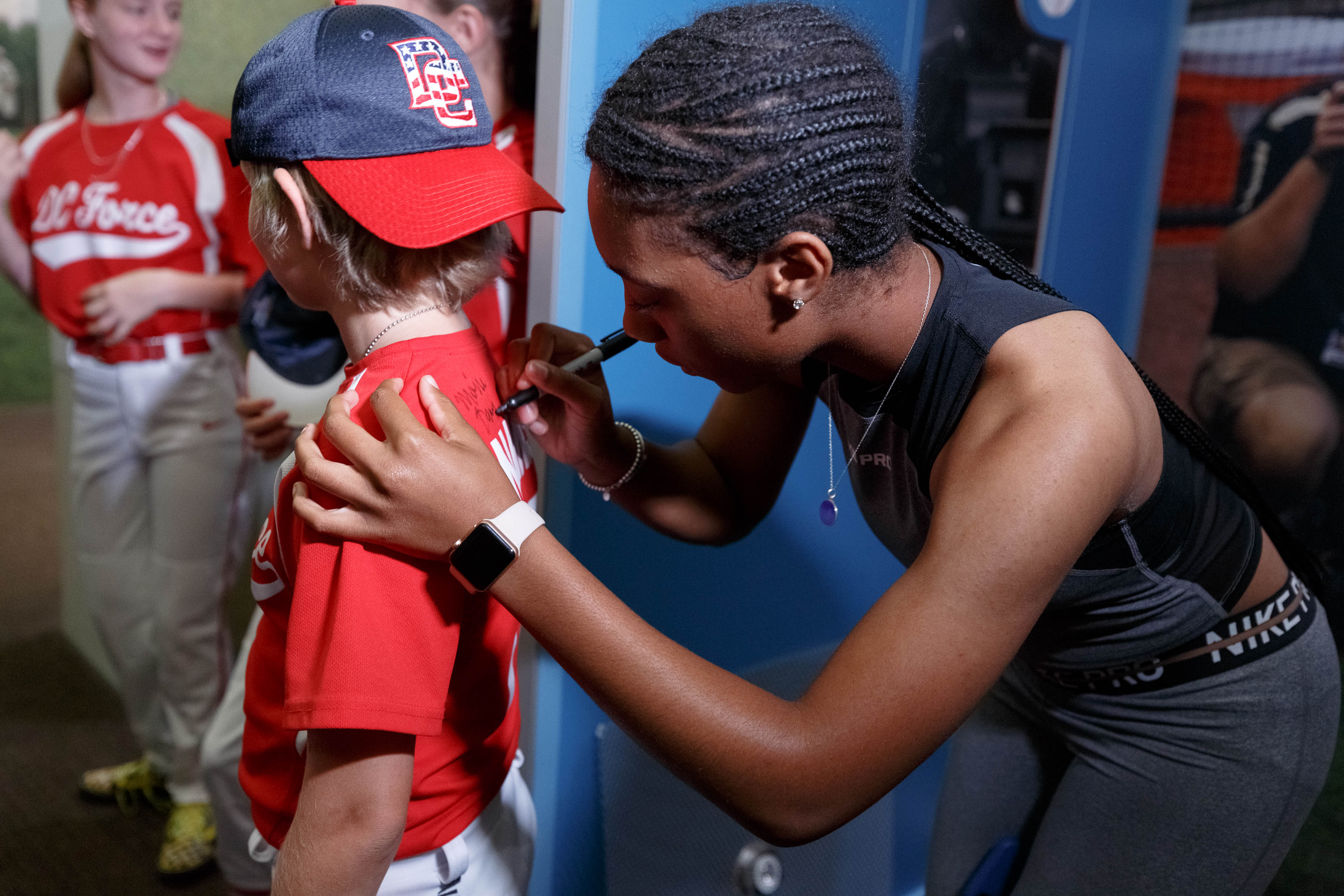 A woman leans down to sign the back of a child's red baseball jersey at an indoor exhibit.