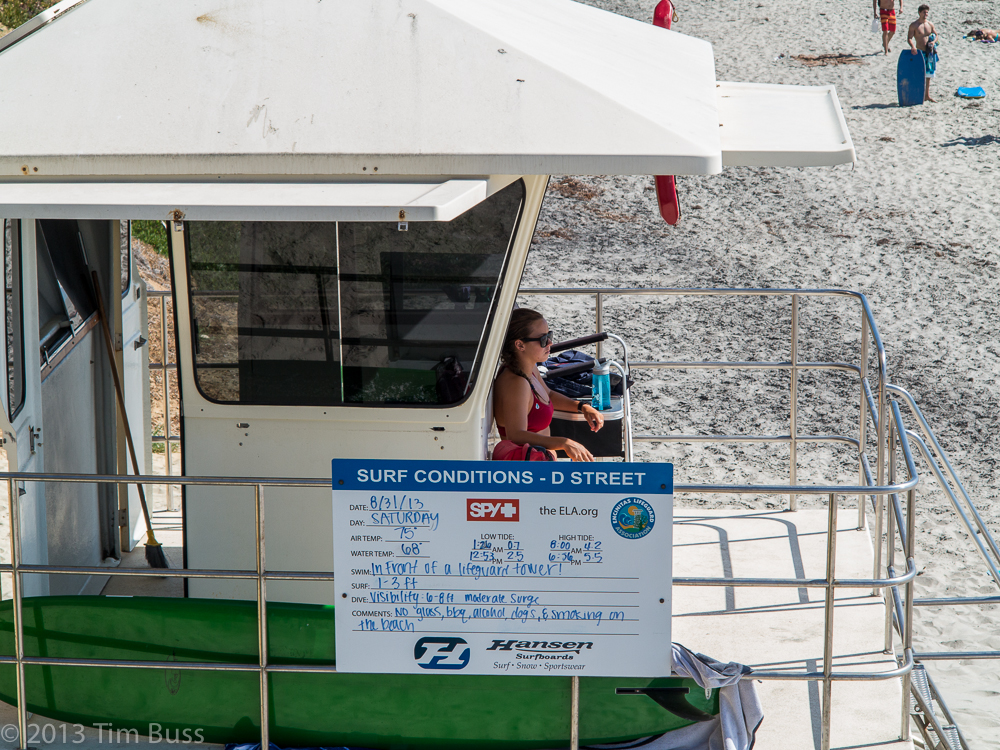 A lifeguard seated inside a beach station booth with a surf conditions sign on the exterior.