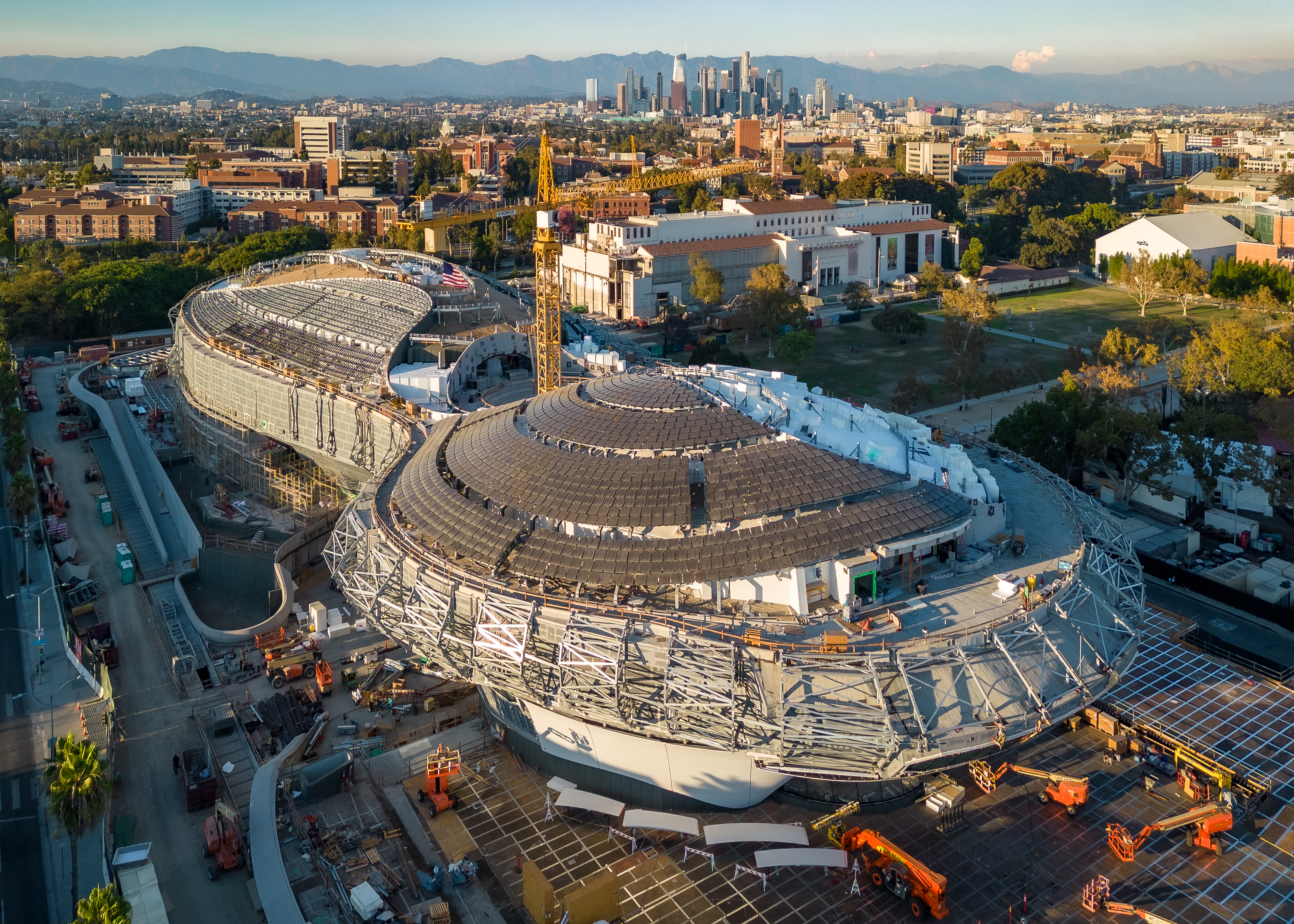 Aerial view of a curved museum building under construction with a city skyline behind it.