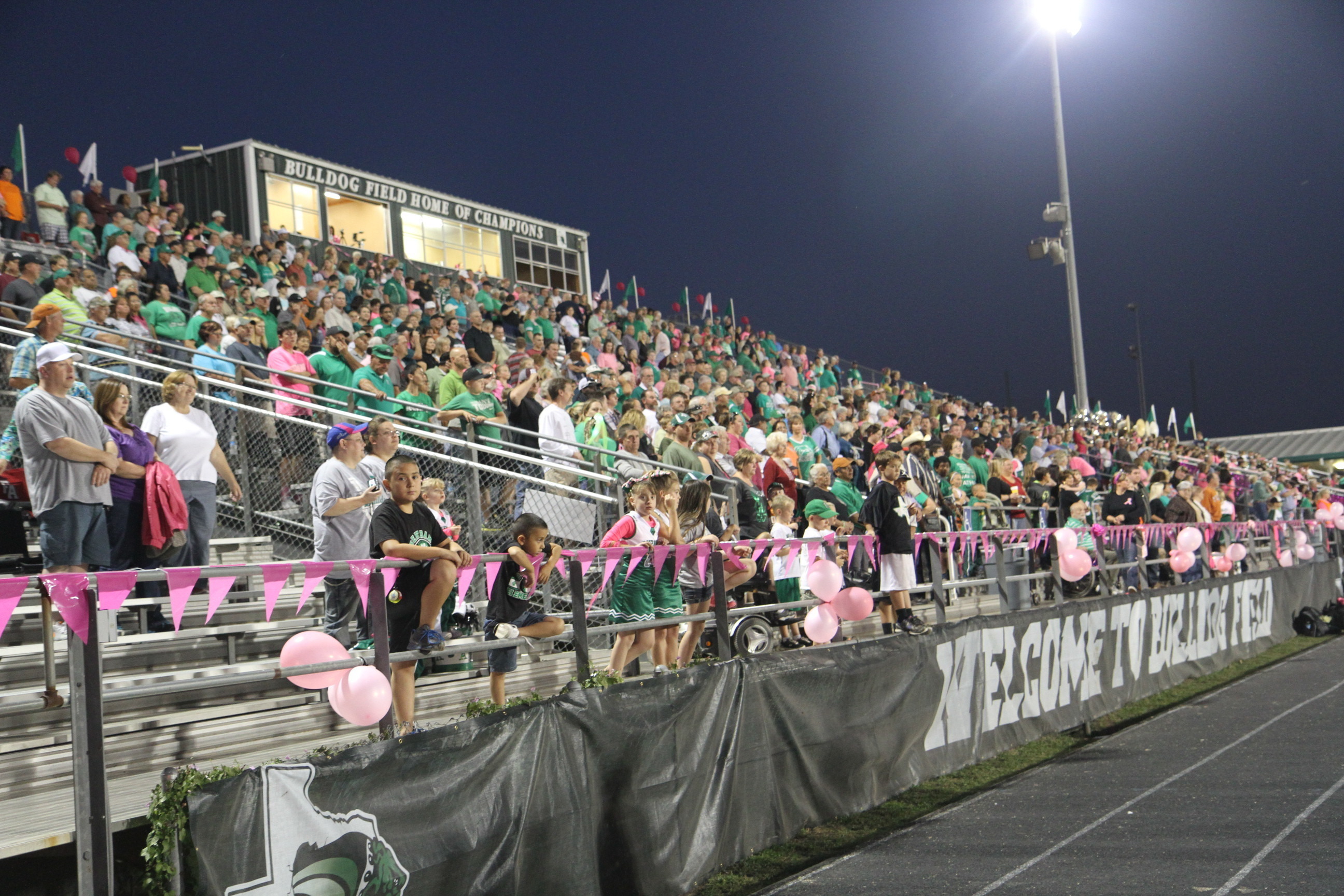 Packed high school football stadium bleachers at night with spectators and pink decorations along the railing.
