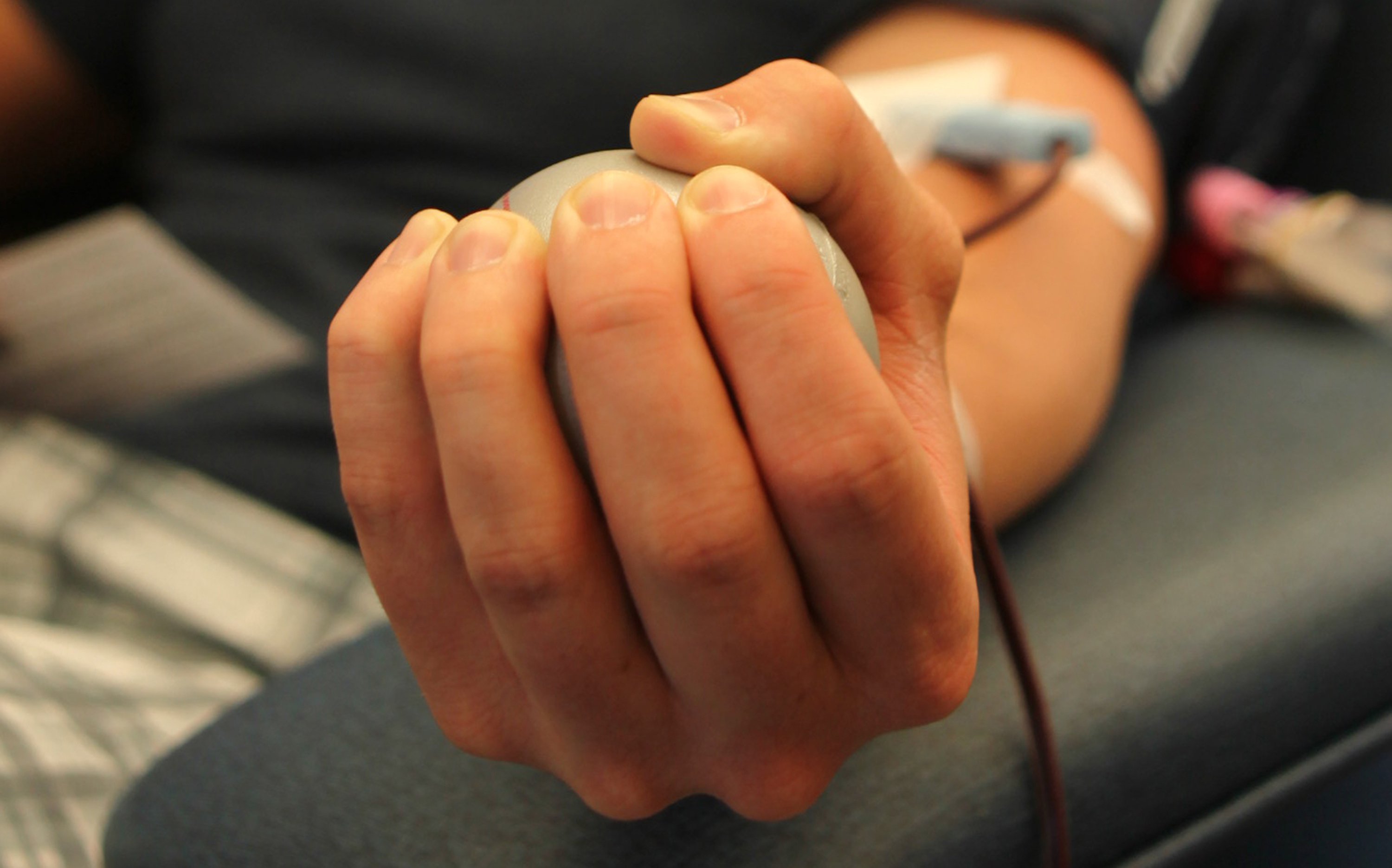 A donor's hand squeezes a stress ball while an IV line draws blood from the forearm.