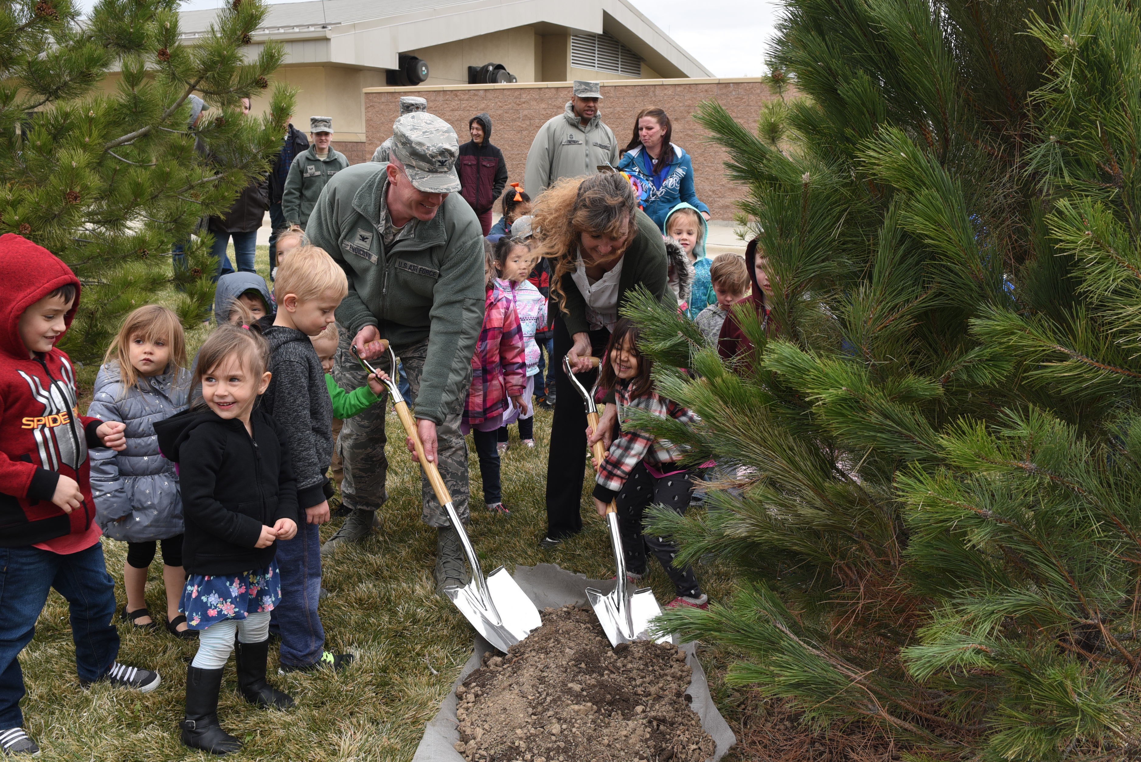 Adults and children using small shovels to plant a pine tree at an outdoor ceremony.