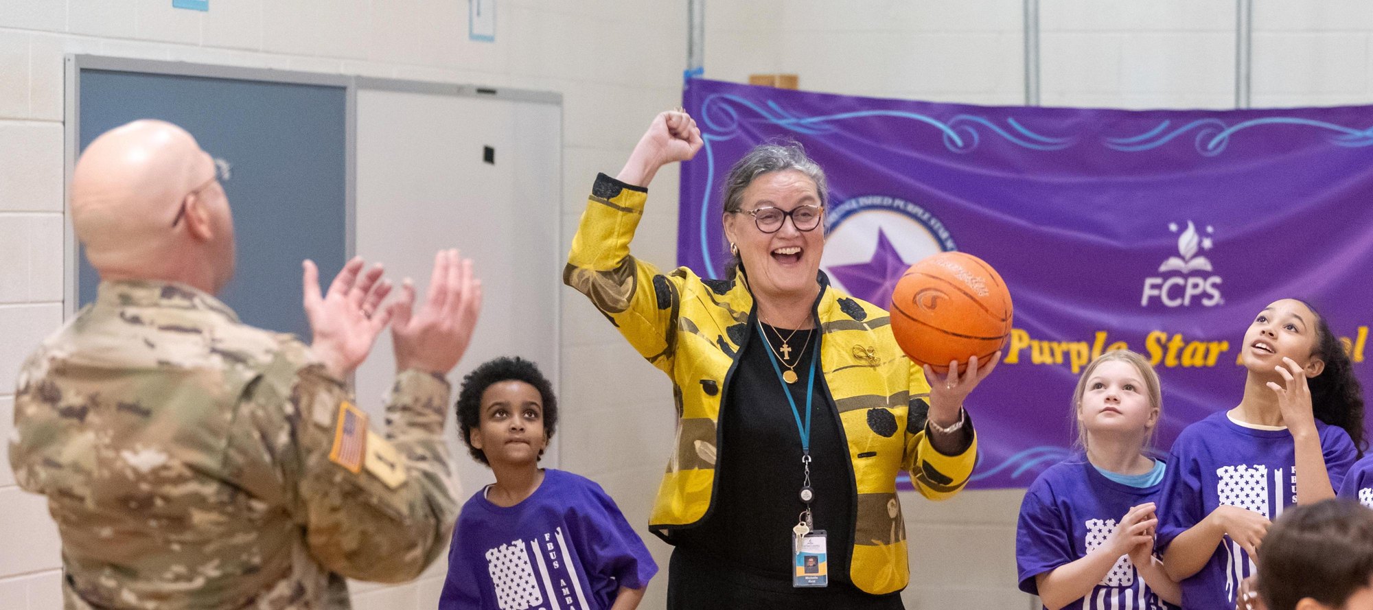 Woman in yellow jacket cheering with children in purple shirts near an FCPS Purple Star banner.