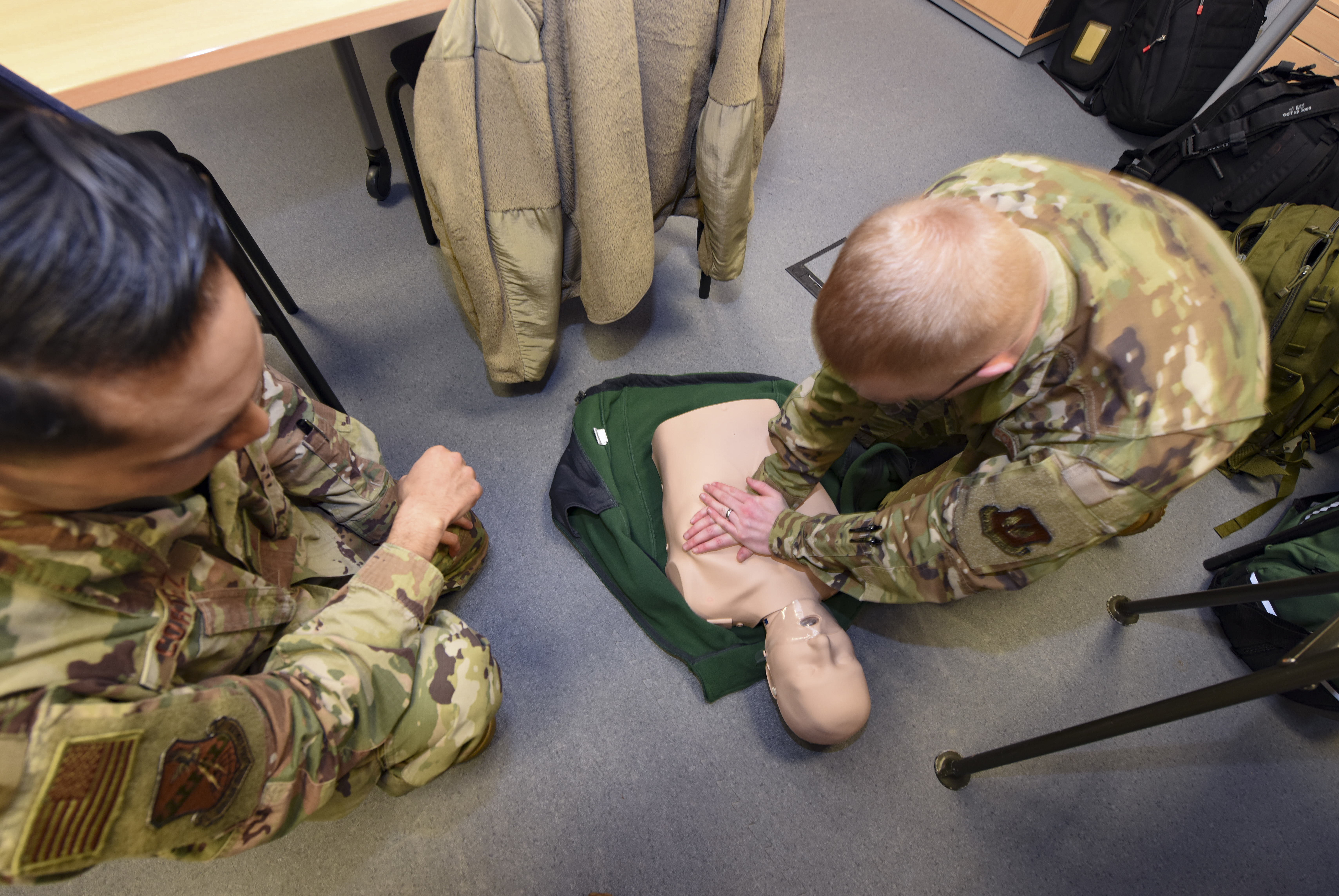 Two people in uniform kneel on the floor practicing chest compressions on a CPR training mannequin.