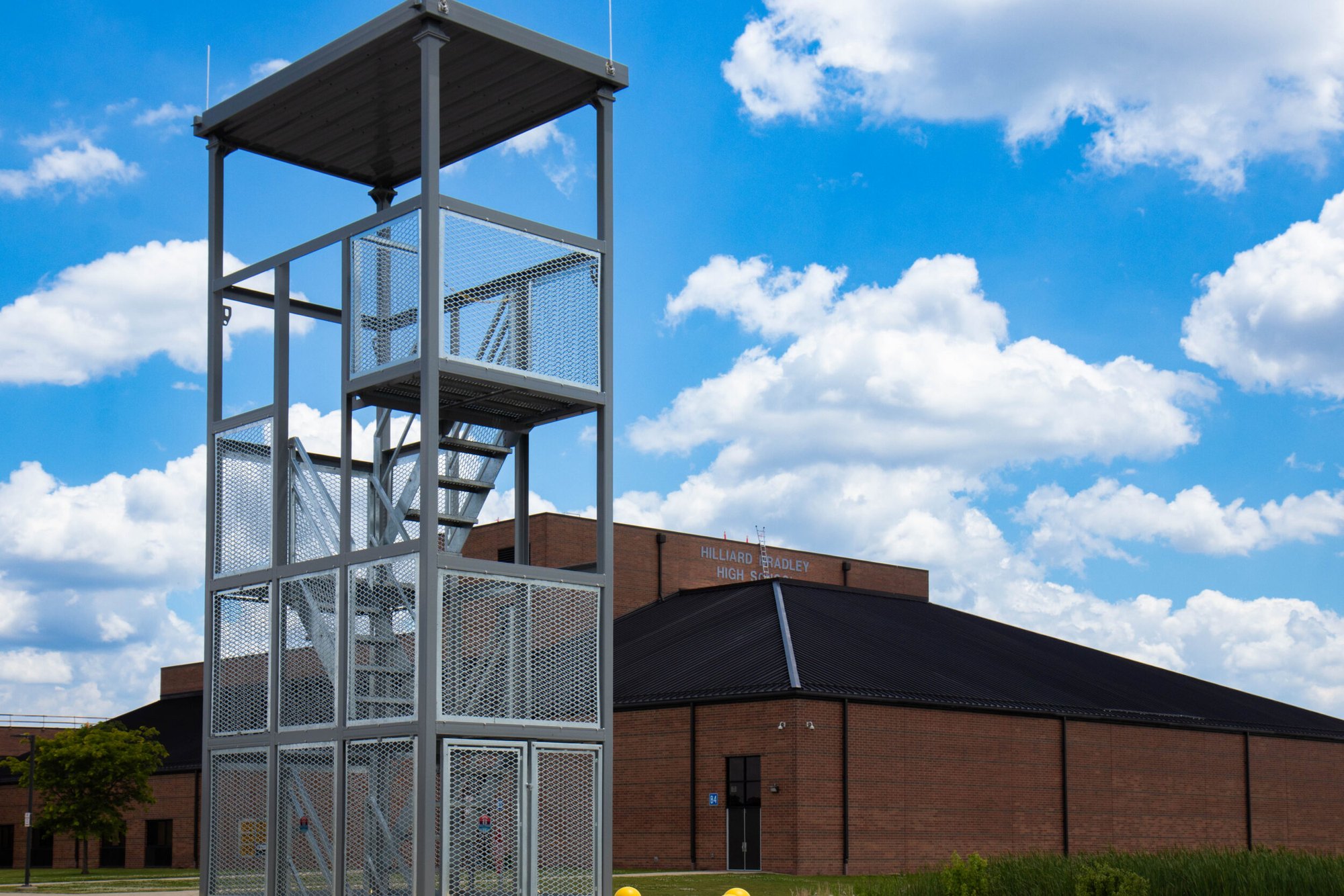 Tall steel mesh band tower with staircases stands beside a brick school building.