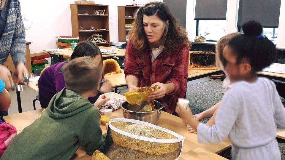 A woman in a red plaid shirt shows honeycomb to children gathered around a table with beekeeping equipment.