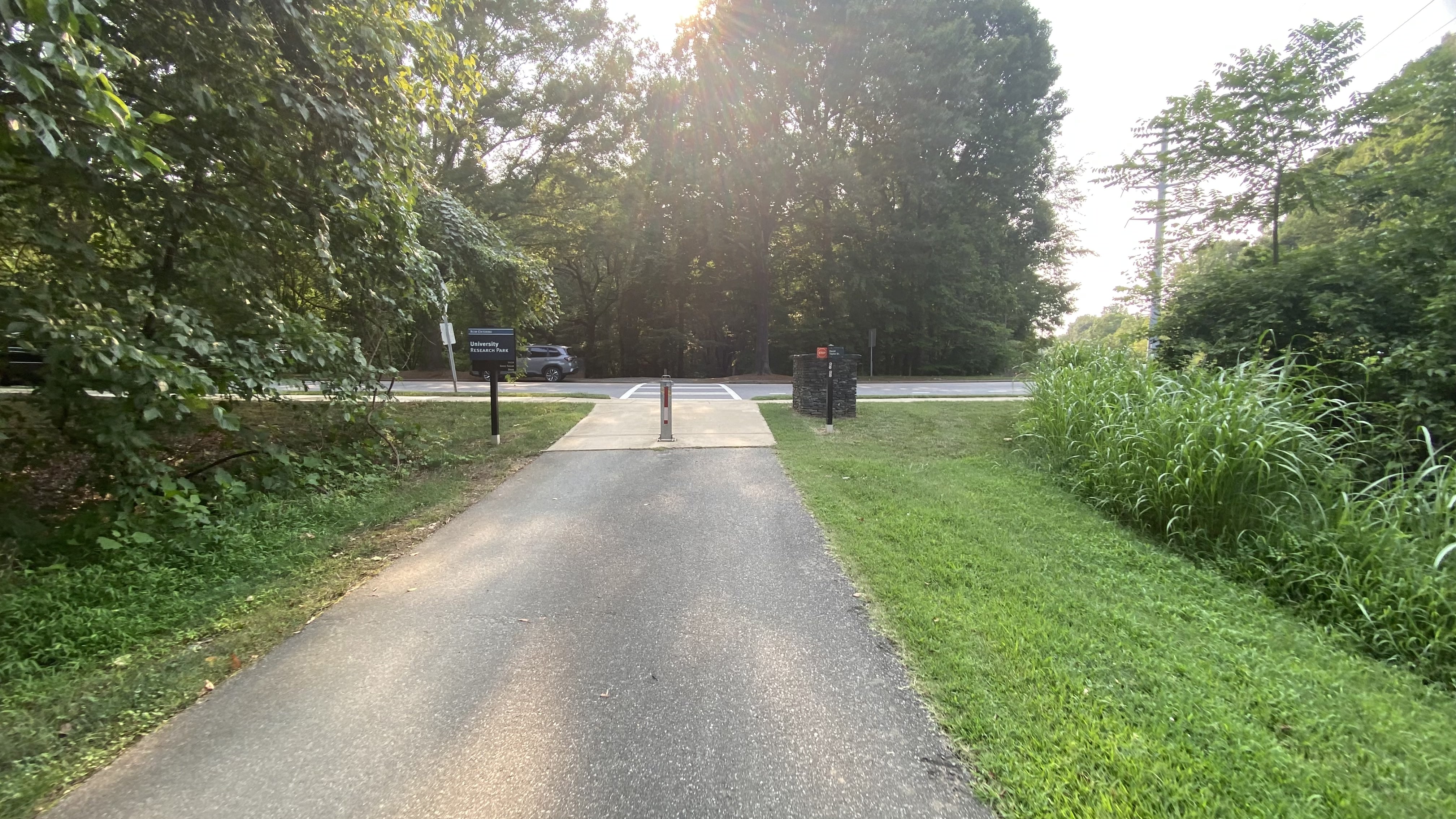 Paved multi-use greenway path lined with trees leading toward a road intersection.