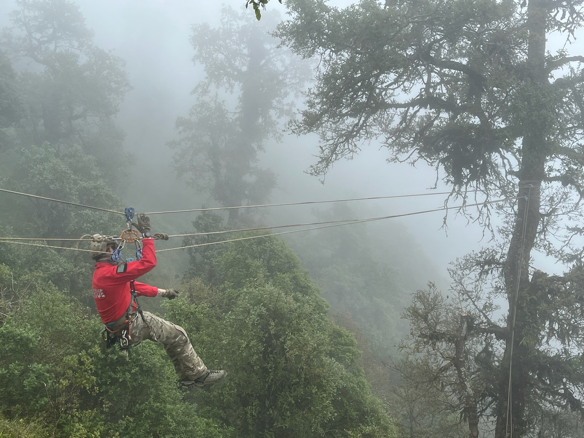 Person in red jacket suspended on a cable through foggy forested mountain terrain.