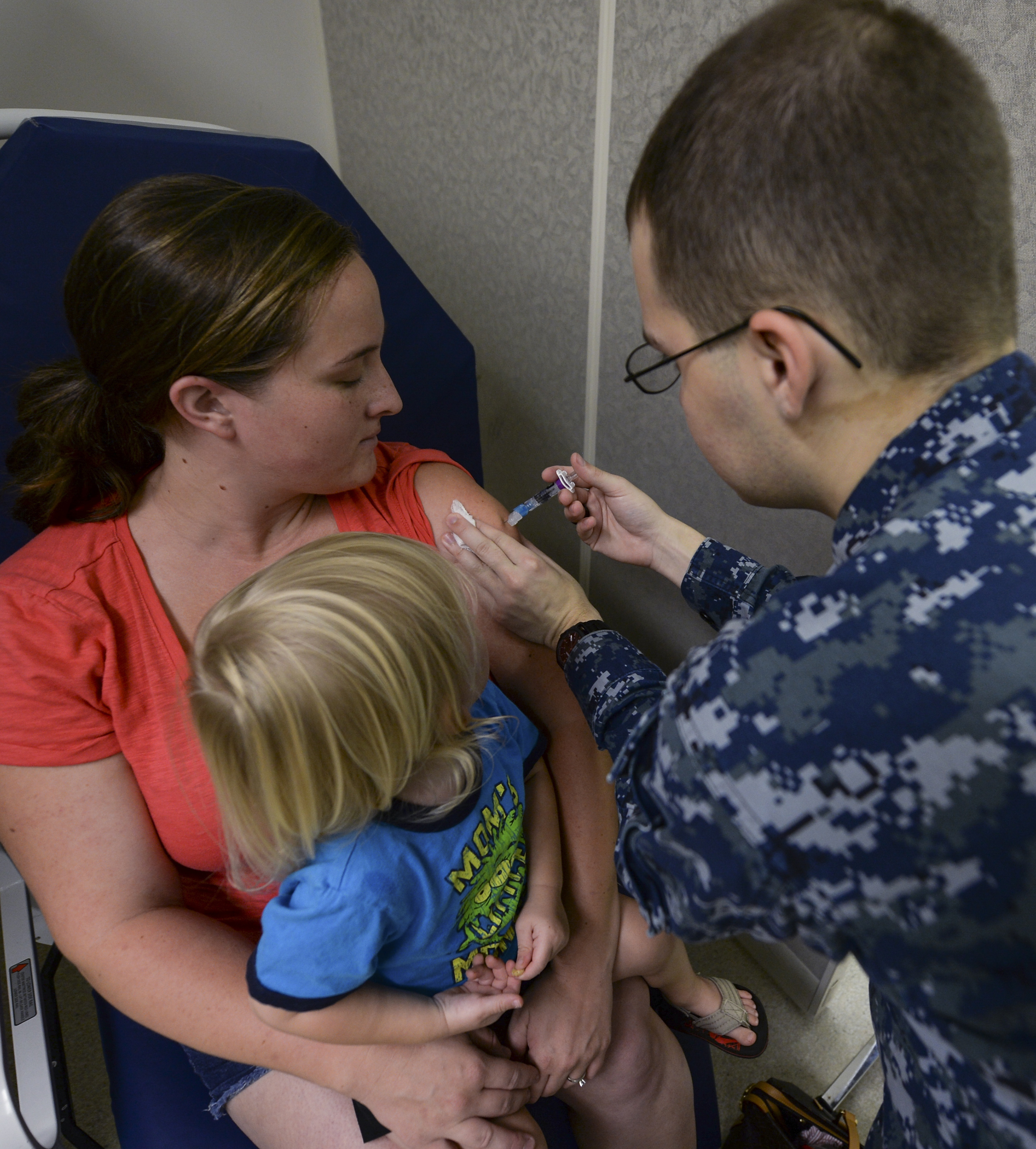 A uniformed healthcare worker gives a flu shot to a woman holding a young child.