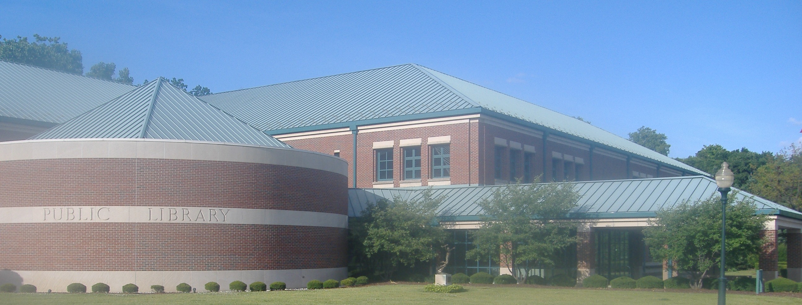 Exterior of a brick public library building with a curved facade and green metal roof.