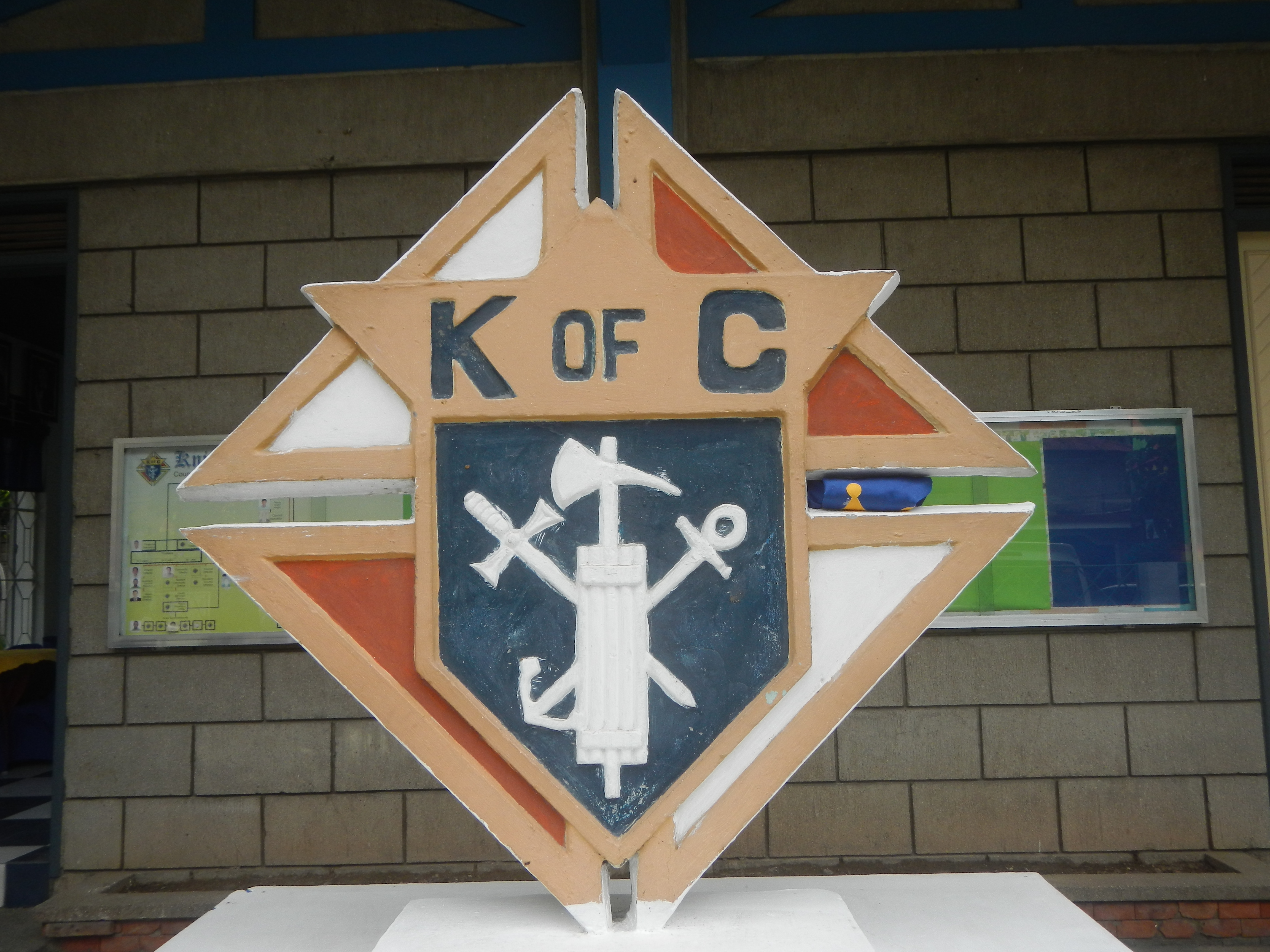 A three-dimensional Knights of Columbus emblem mounted on a white pedestal outdoors.