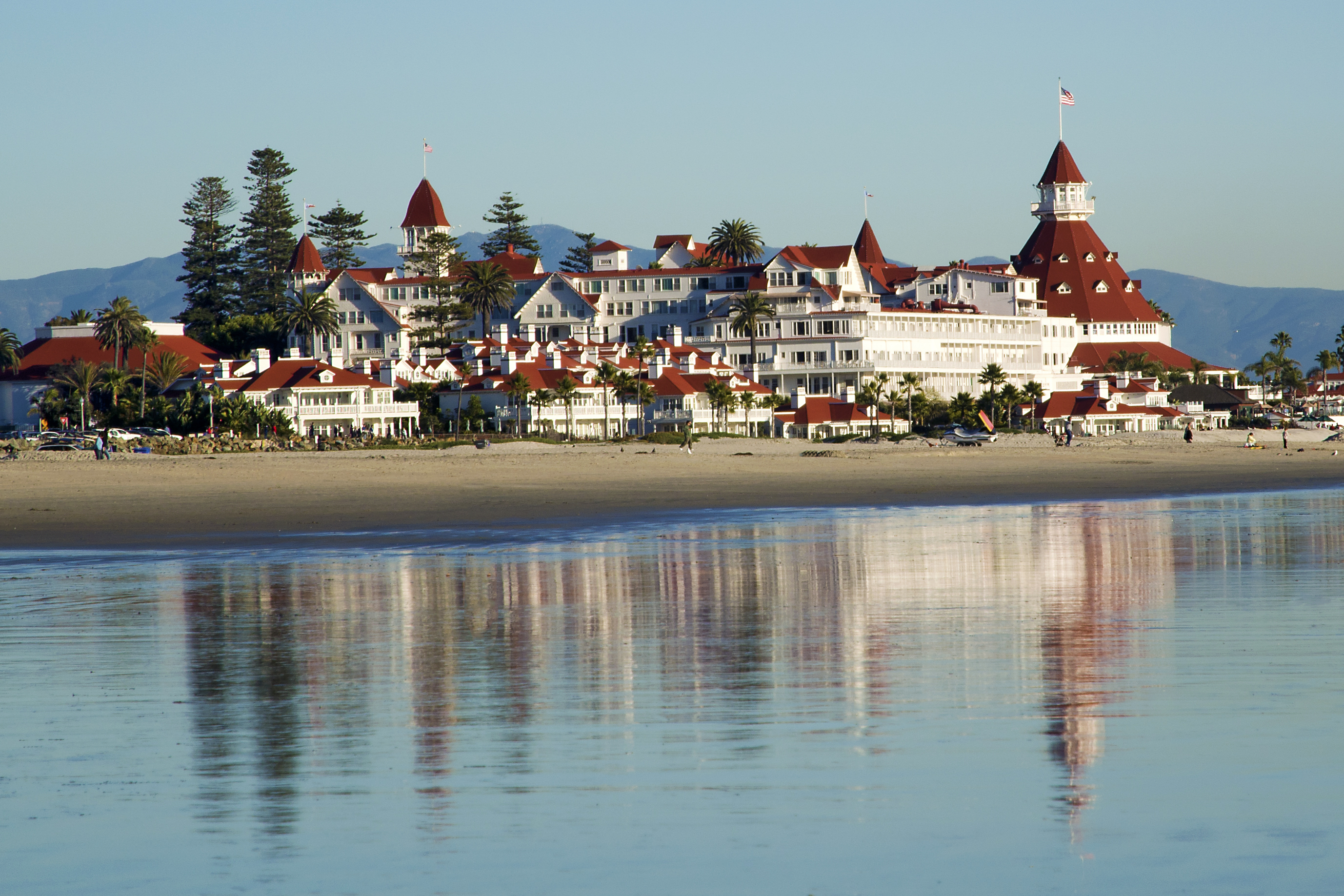 Large Victorian resort hotel with red roofs seen from the beach, shallow water in foreground.