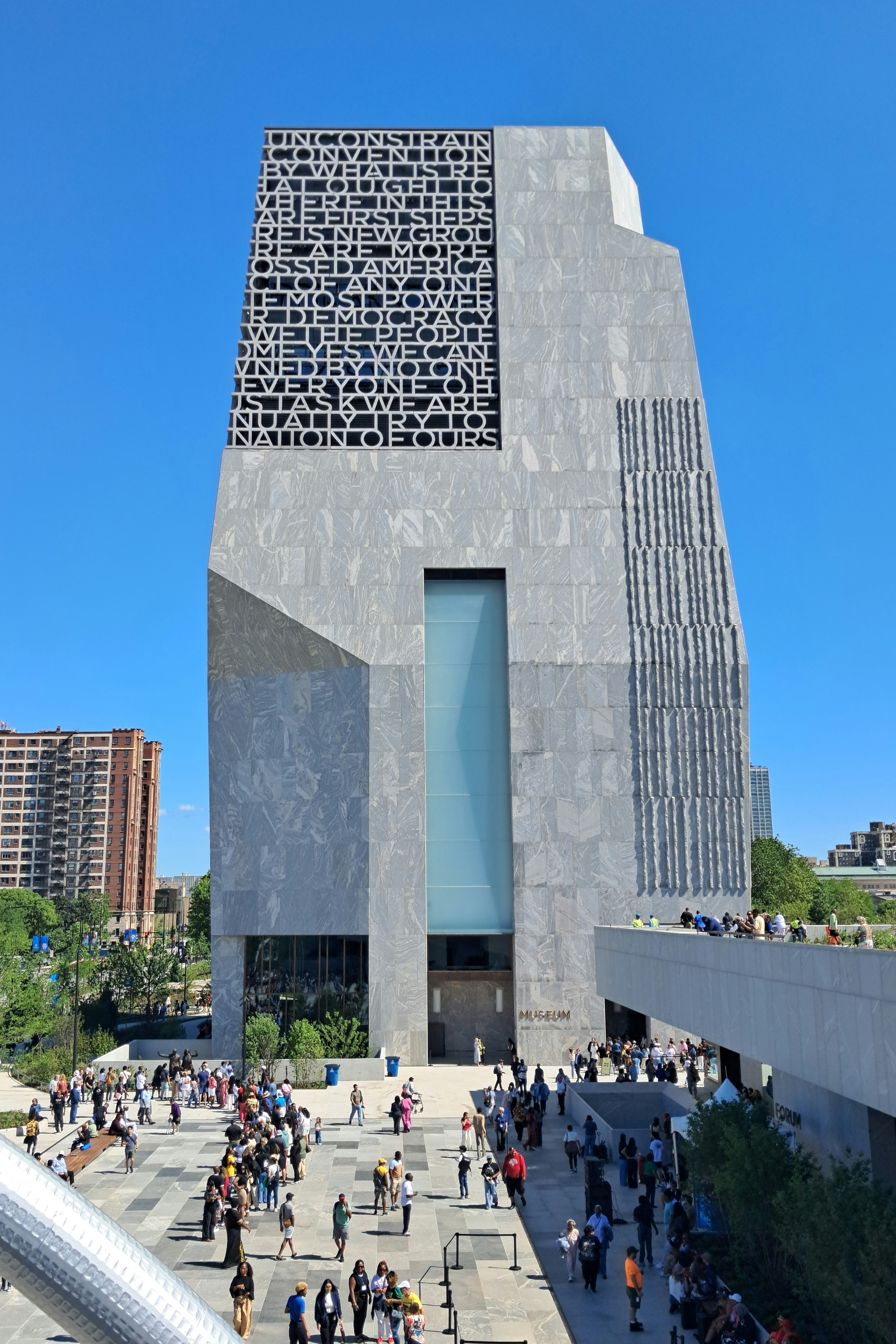 Tall stone tower building with engraved text, surrounded by crowds on a sunny plaza.