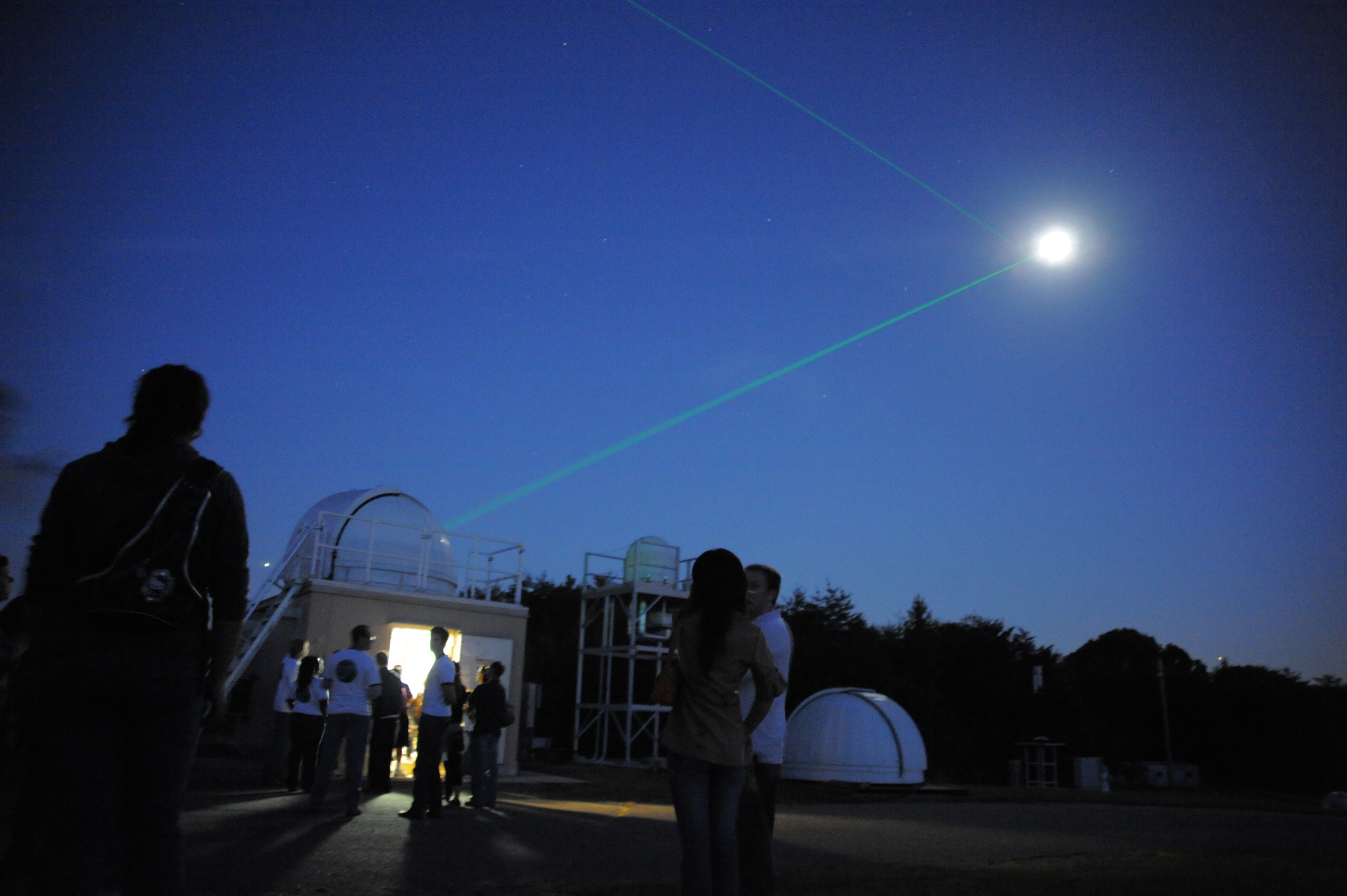 Silhouetted visitors watch a green laser beam aimed at the moon near observatory domes at night.