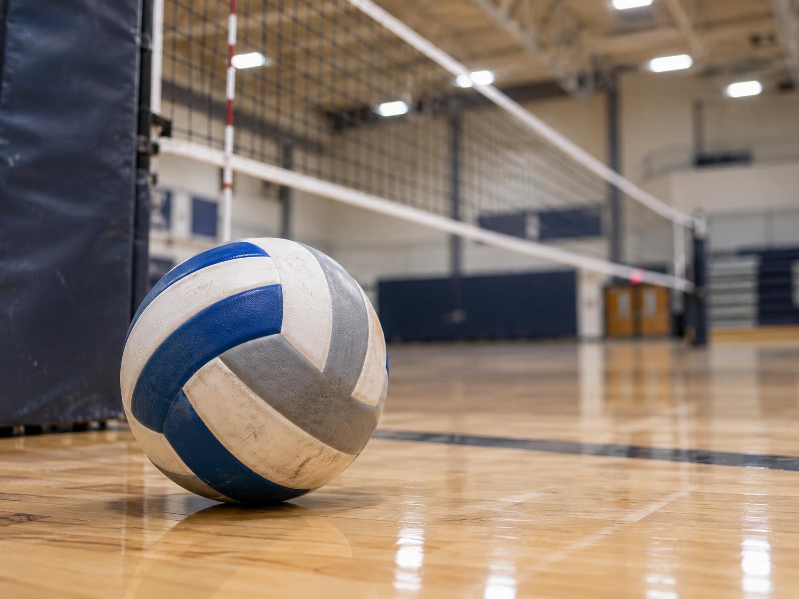 A volleyball resting on a hardwood gymnasium floor near a net post.