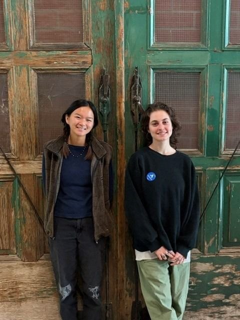 Two smiling young women stand side by side in front of large green wooden doors.