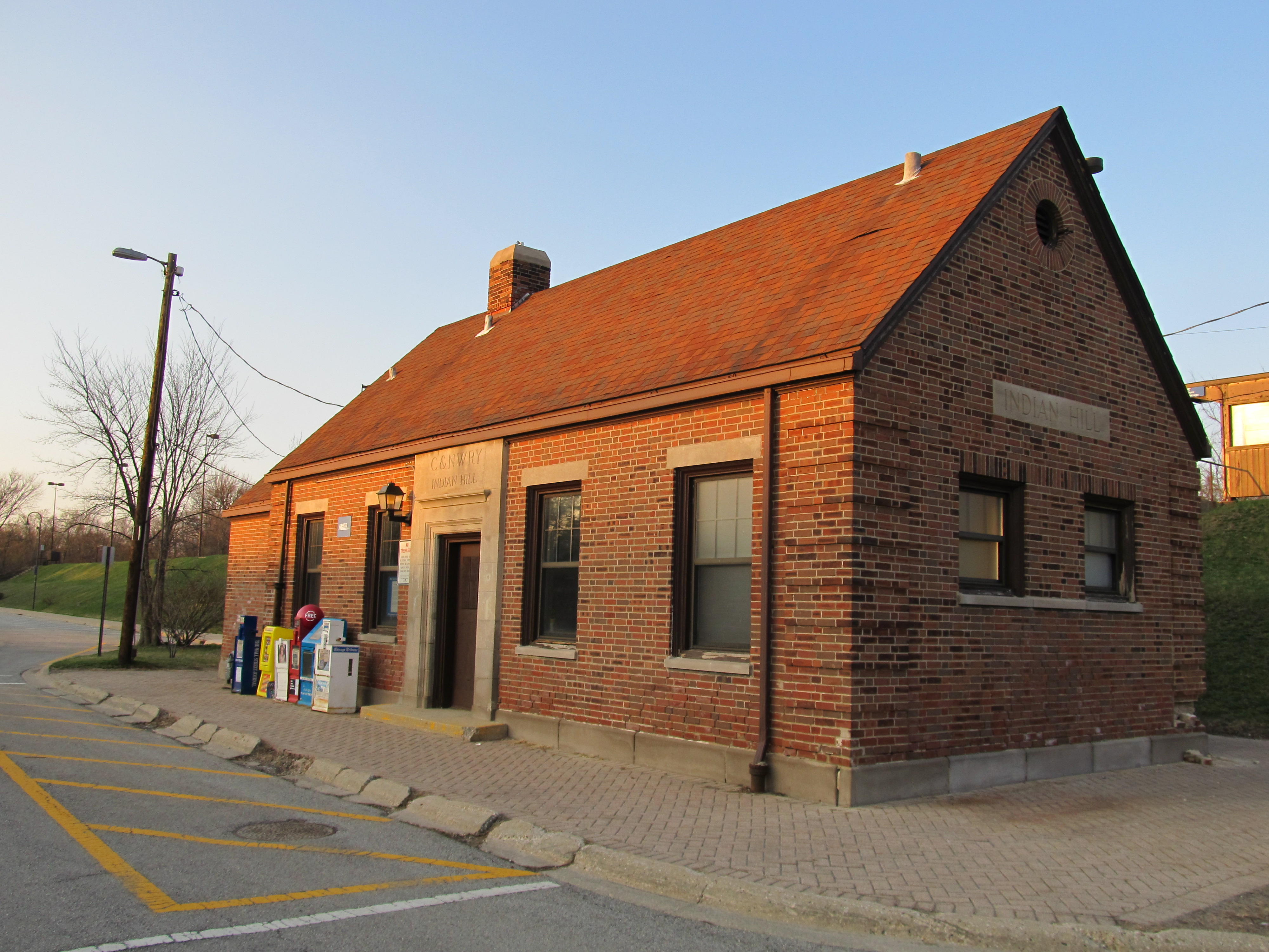 Small red brick Metra station building with a steeply pitched roof in a parking lot.