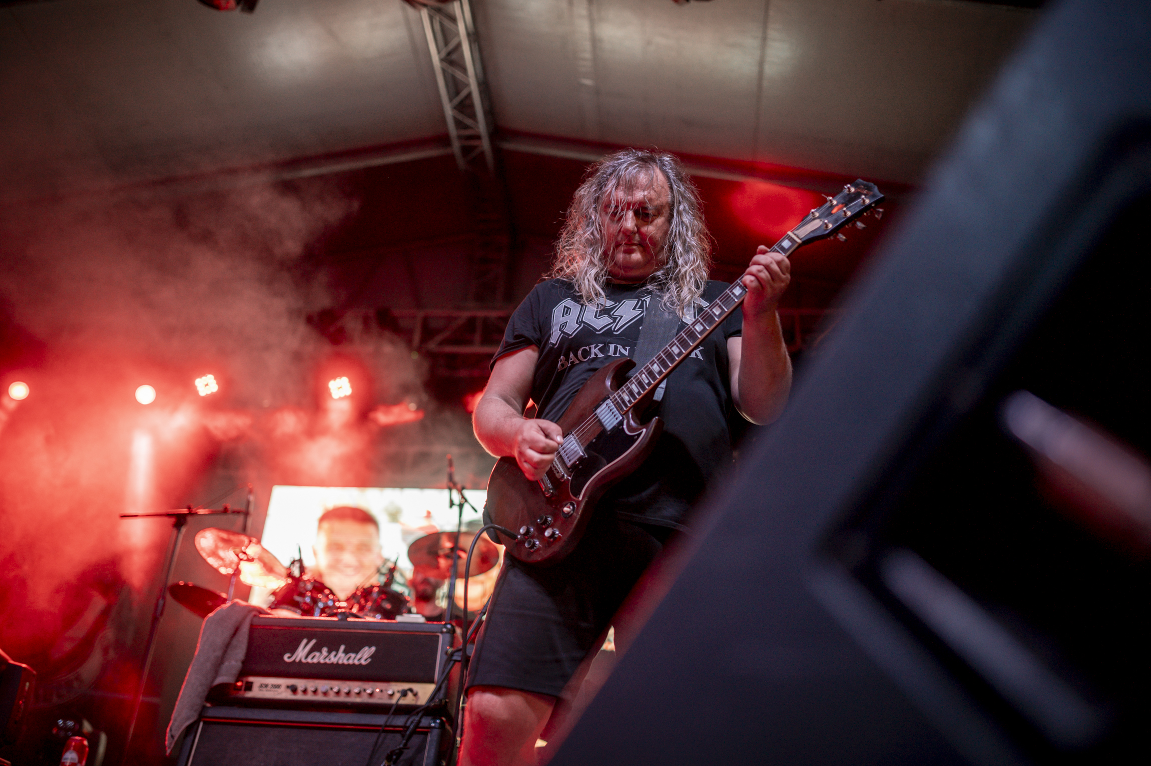 Guitarist in black t-shirt plays electric guitar on a brightly lit outdoor stage.