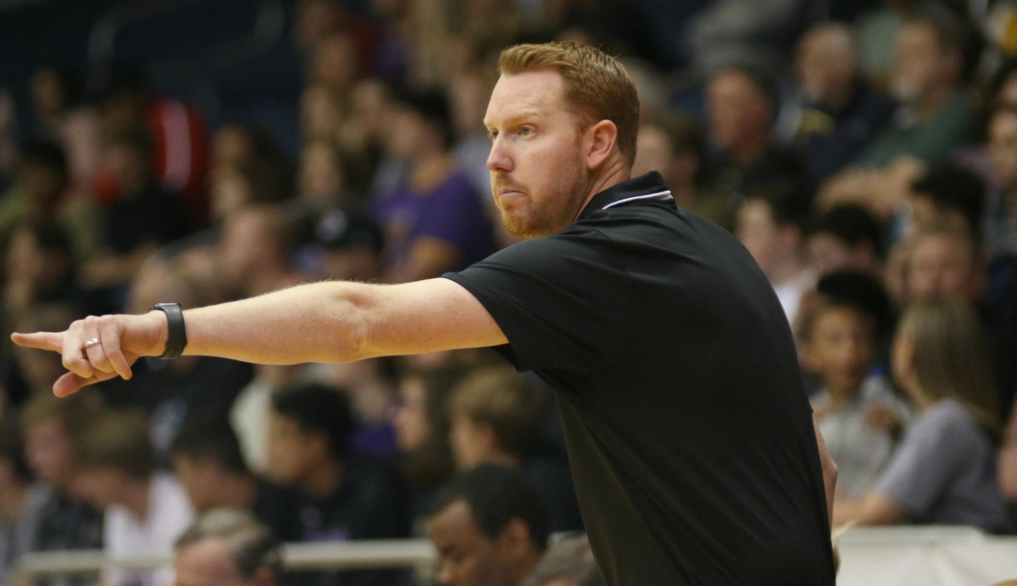 A red-bearded man in a black shirt gestures on a gymnasium sideline with bleachers behind him.