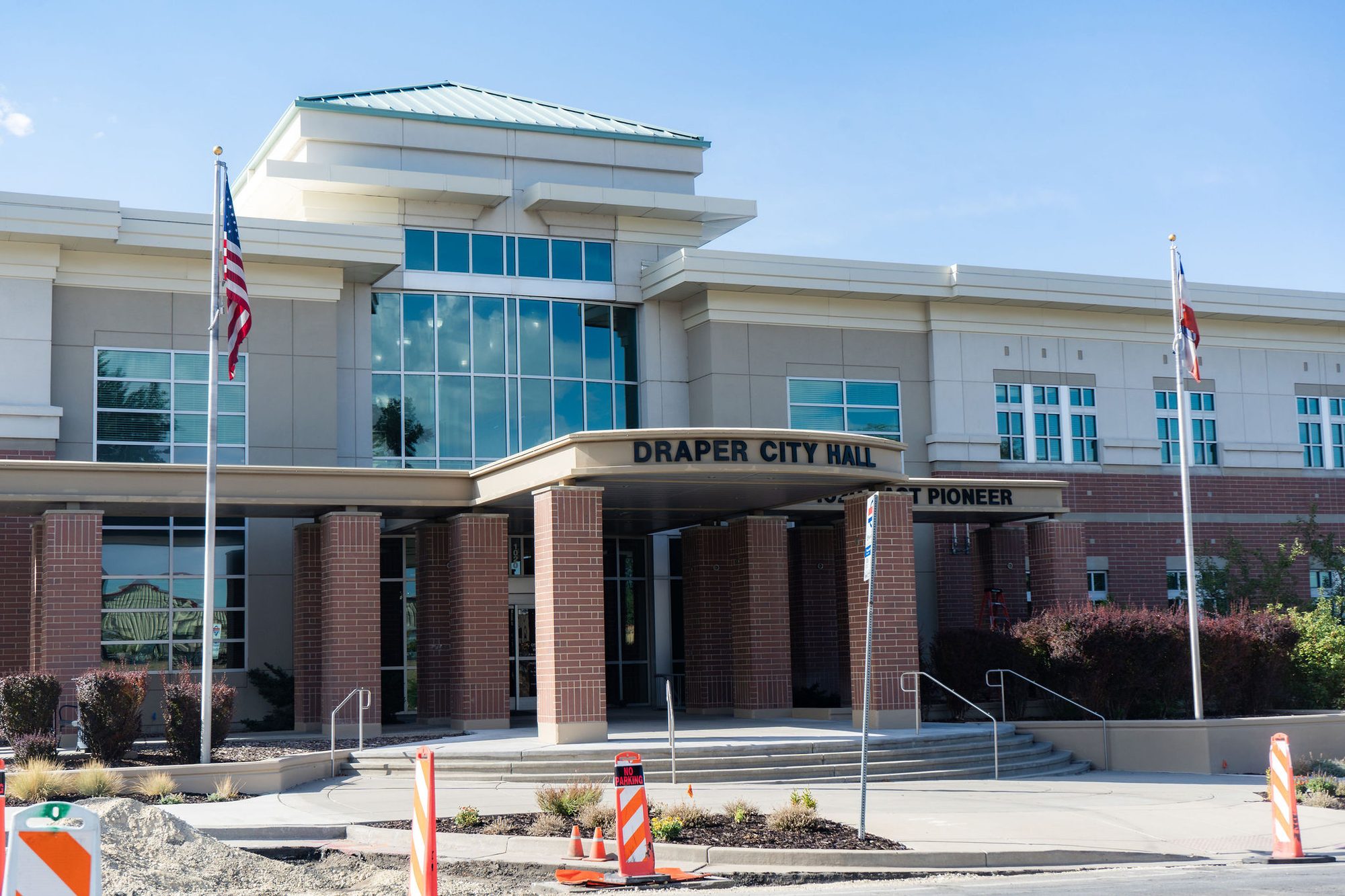 Exterior of a two-story brick city hall building with flags and orange construction cones in front.