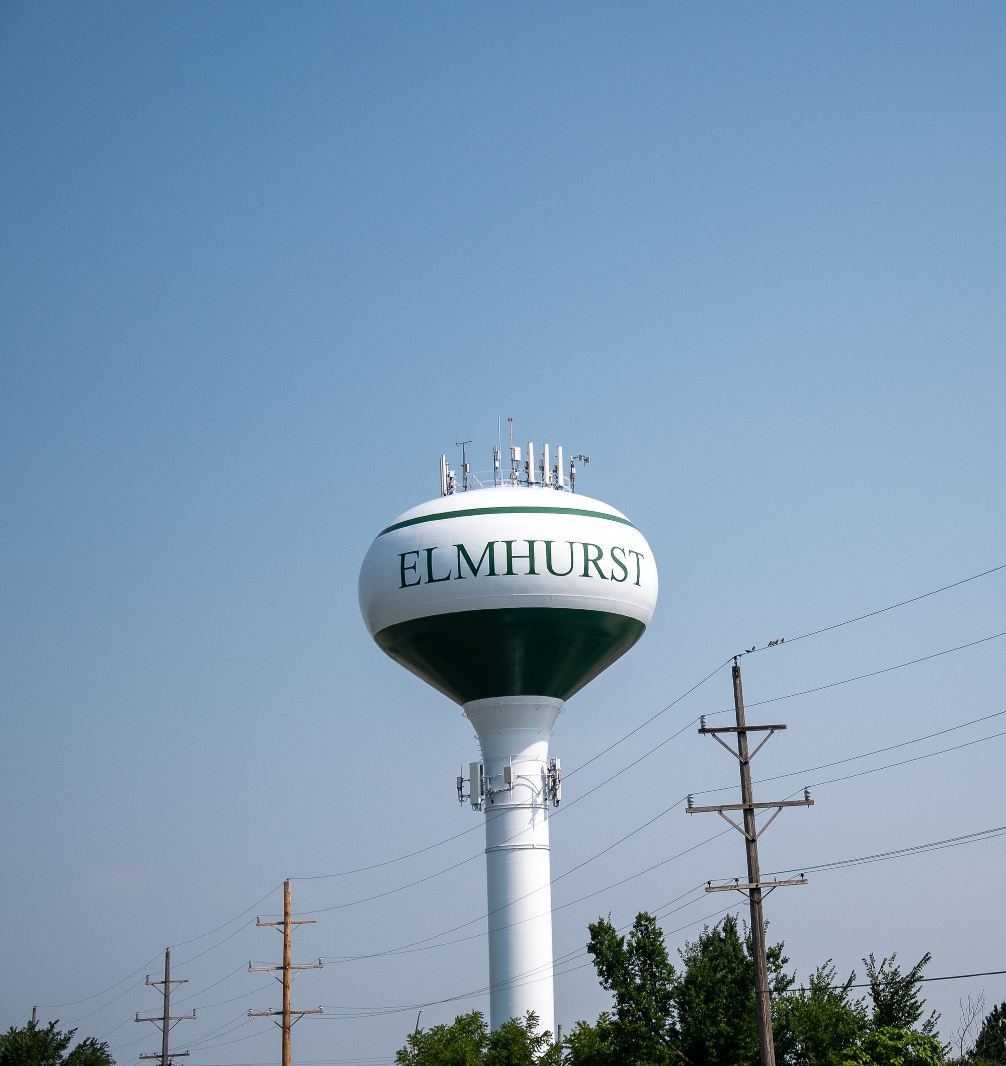 White municipal water tower with green lettering rising above utility poles and trees.