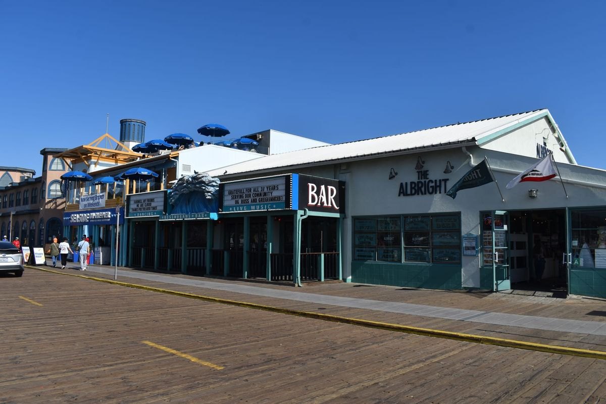 Wooden boardwalk on Santa Monica Pier with restaurant buildings and blue umbrellas on a rooftop deck.