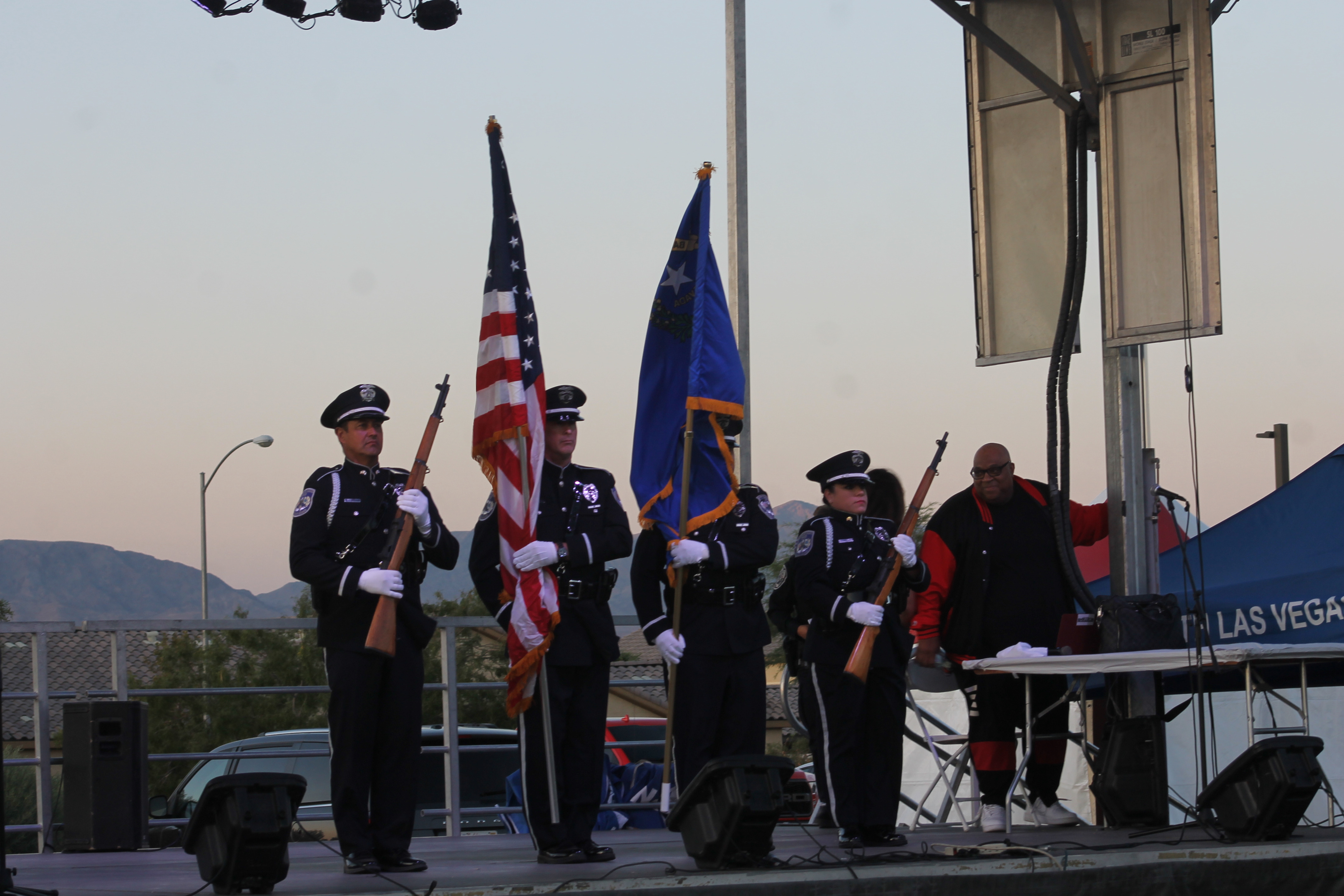Police officers in a color guard formation hold rifles and flags on an outdoor stage at dusk.