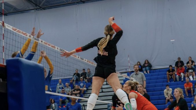 Female volleyball player in black jersey jumping to spike ball over the net.
