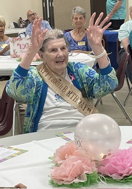 Elderly woman in gold birthday sash and tiara raises both hands at a decorated party table.