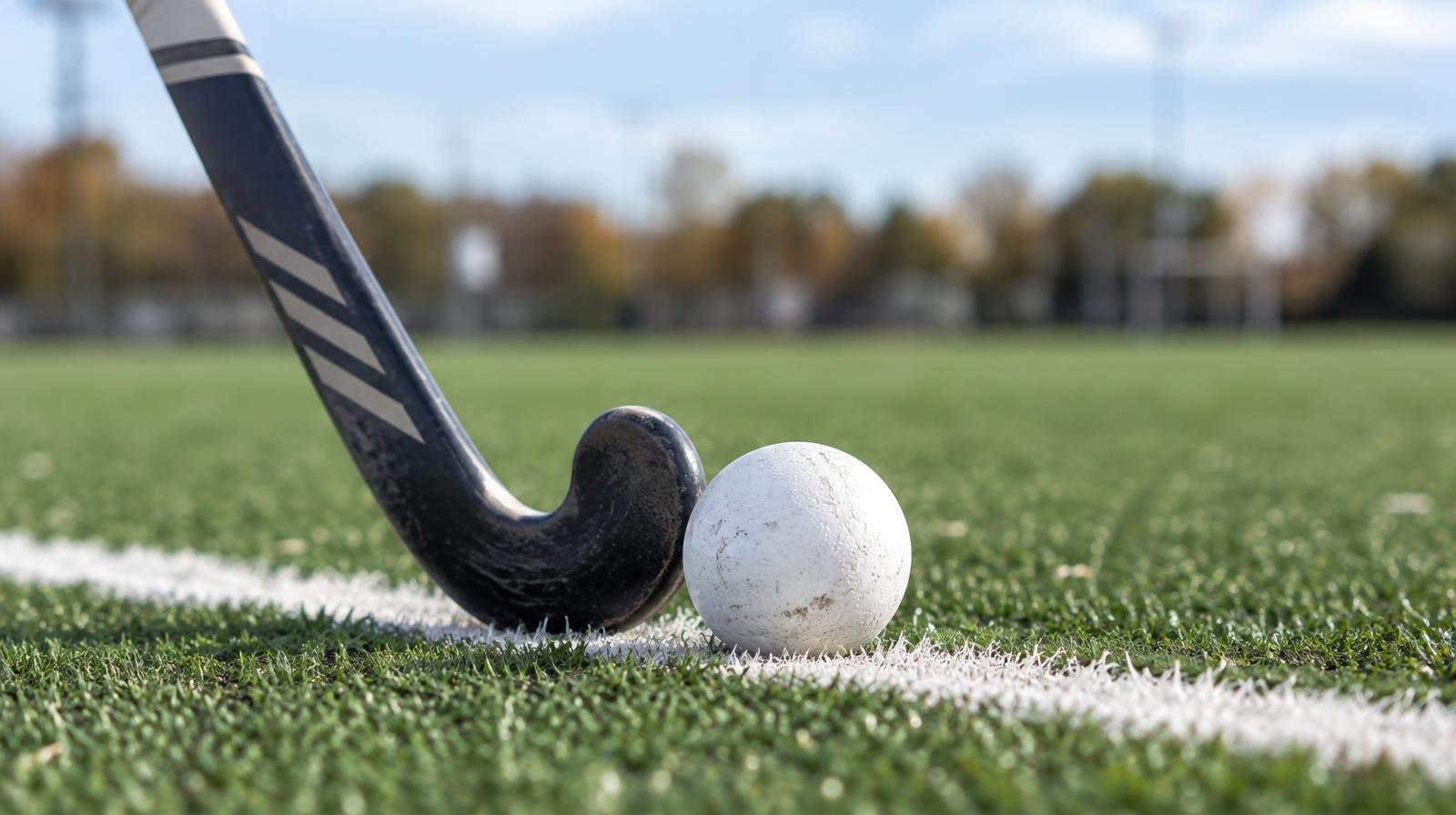 A field hockey stick and orange ball resting on green artificial turf beside a white line.