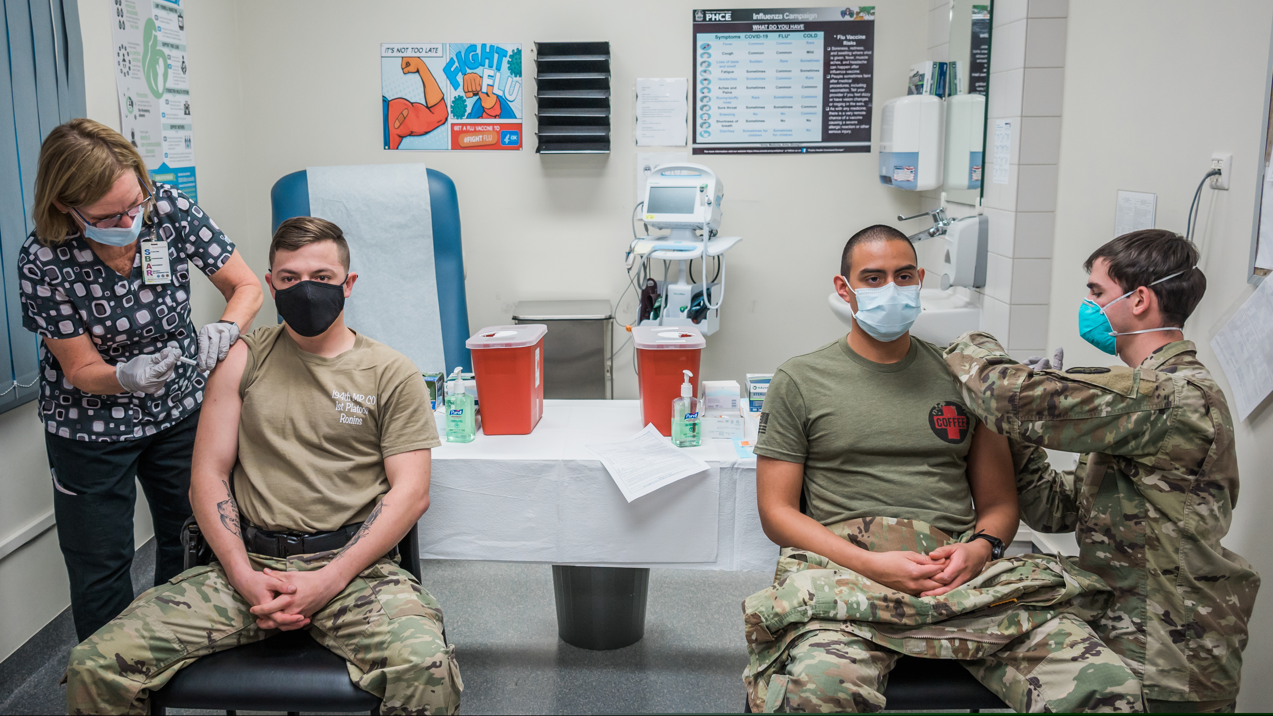 Two people receiving vaccine injections simultaneously from masked healthcare workers in a clinic.