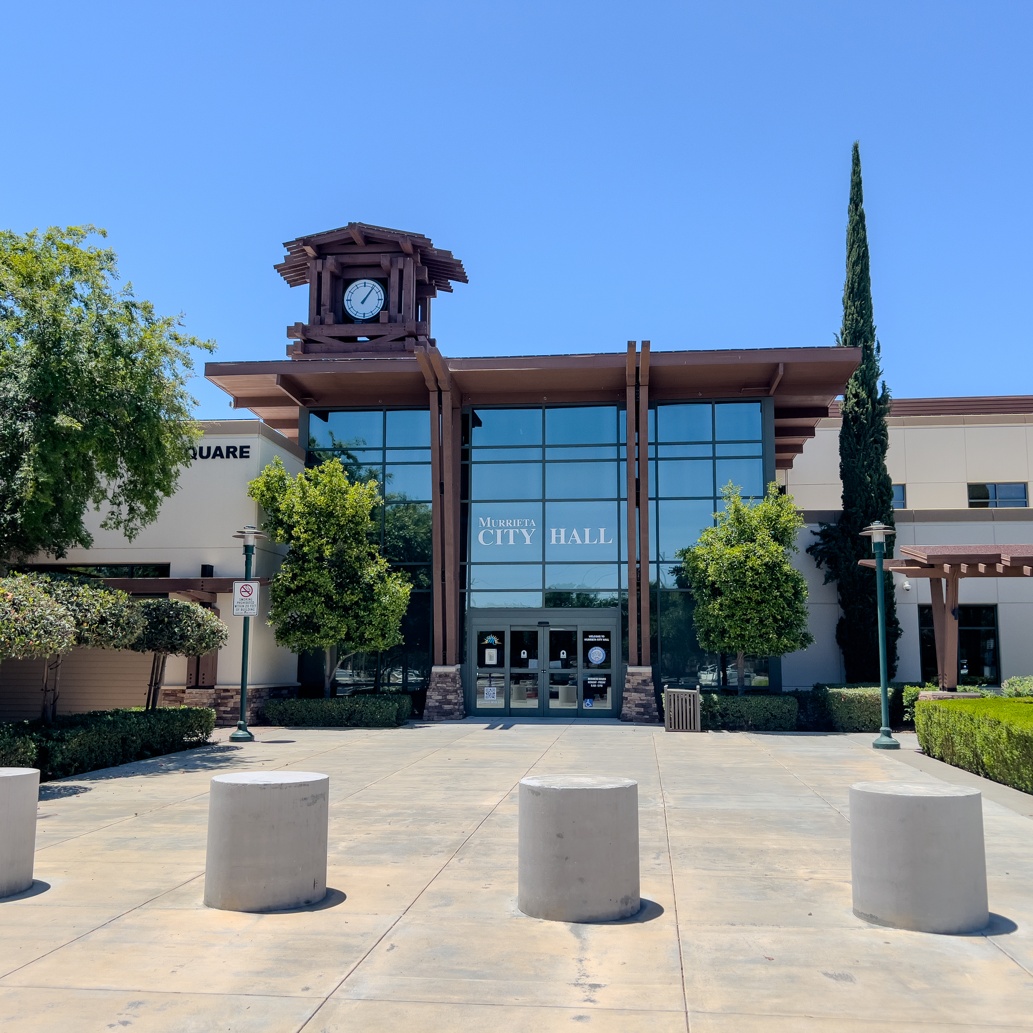 Exterior of a city hall building with a clock tower, glass entrance, and cypress trees.