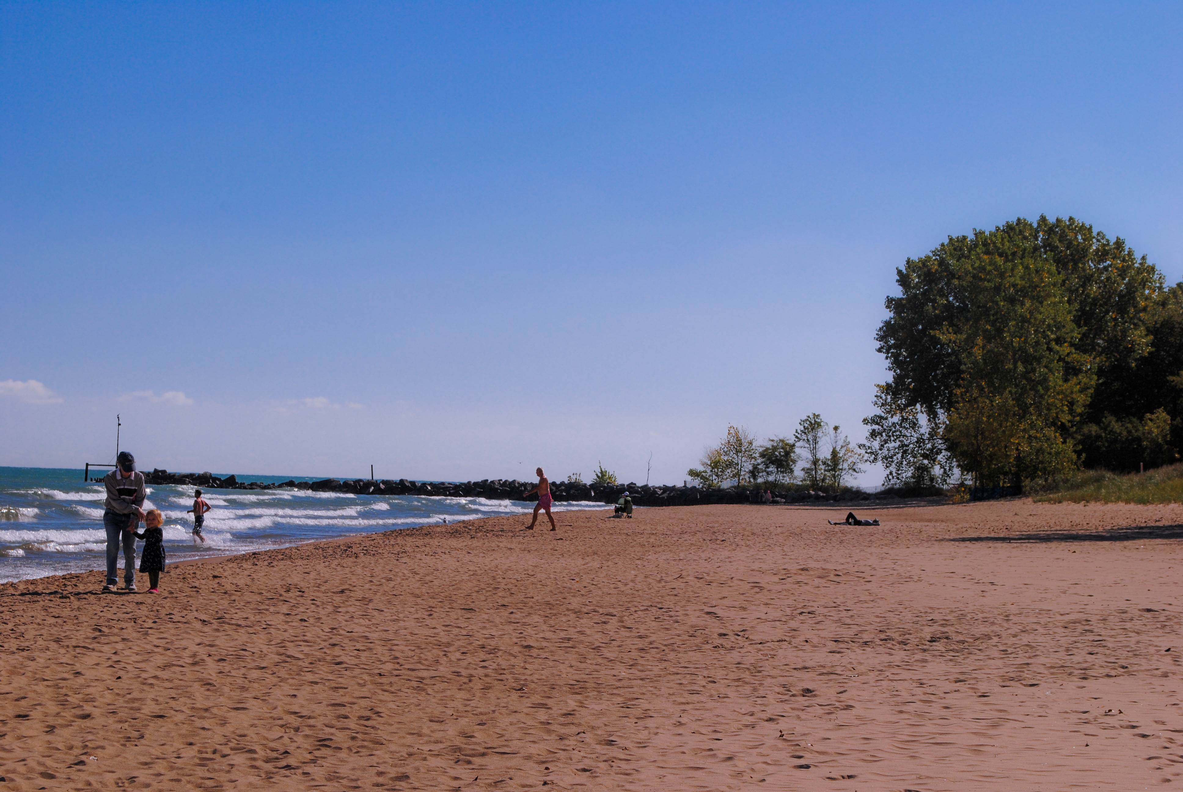 Sandy beach along Lake Michigan with beachgoers, a rocky breakwater, and green trees under a blue sky.