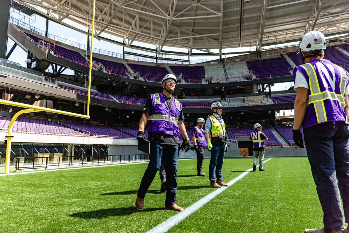 Construction workers in hard hats stand on a turf field inside a stadium with purple seats.