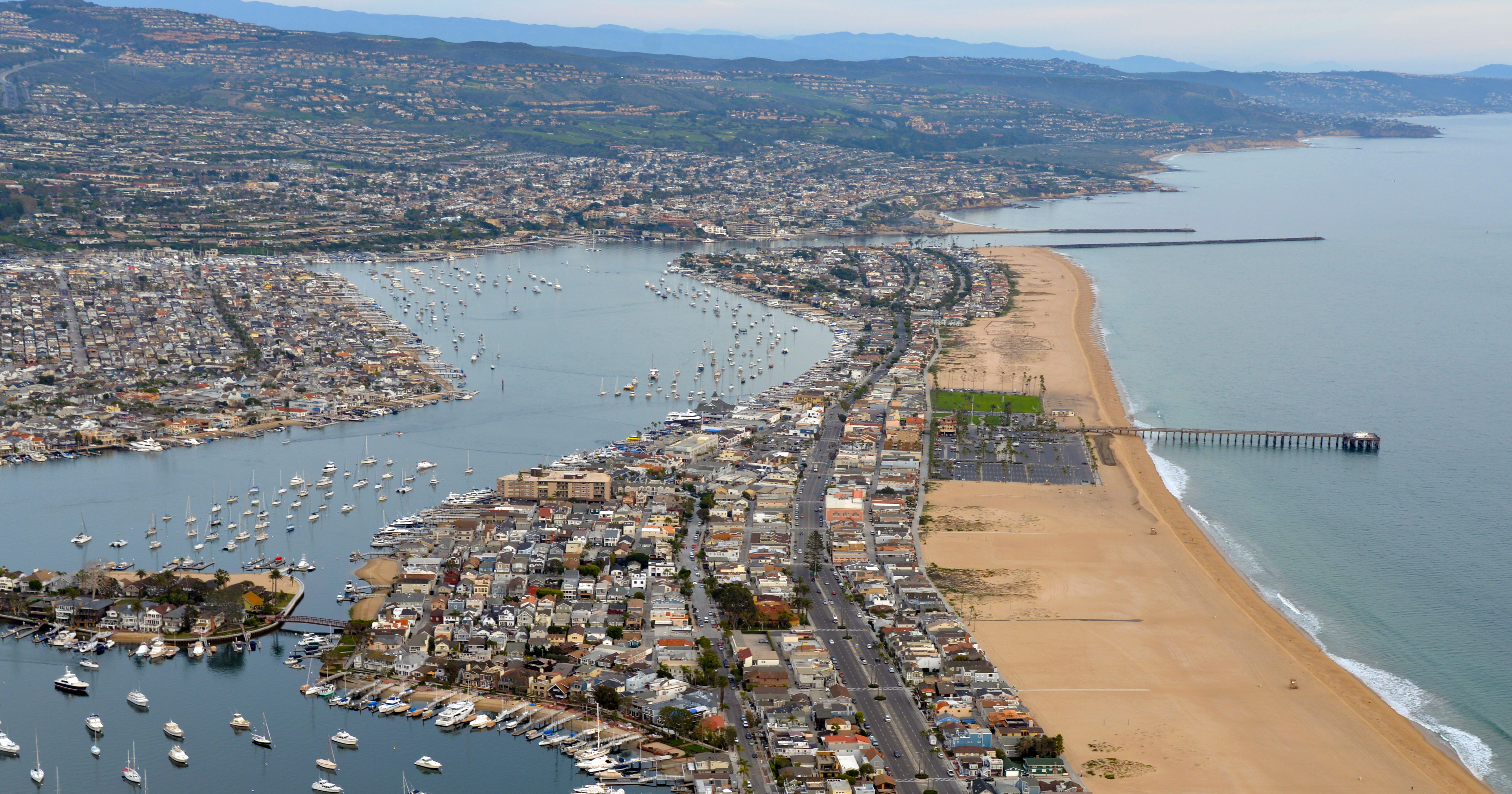 Aerial view of a narrow coastal peninsula with a pier, beach, and sailboat-filled bay.