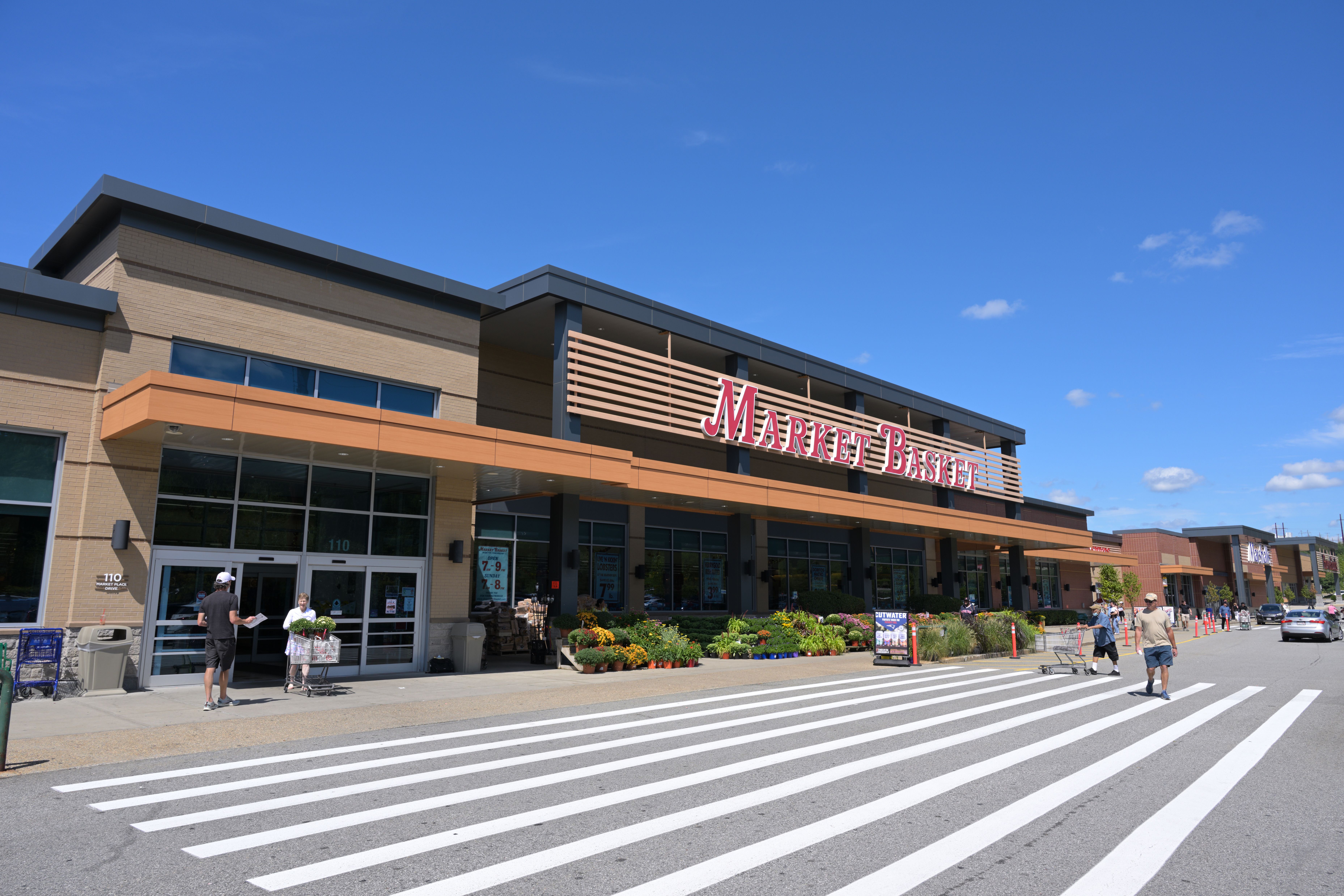 Exterior of a Market Basket supermarket with red signage and a parking lot on a sunny day.