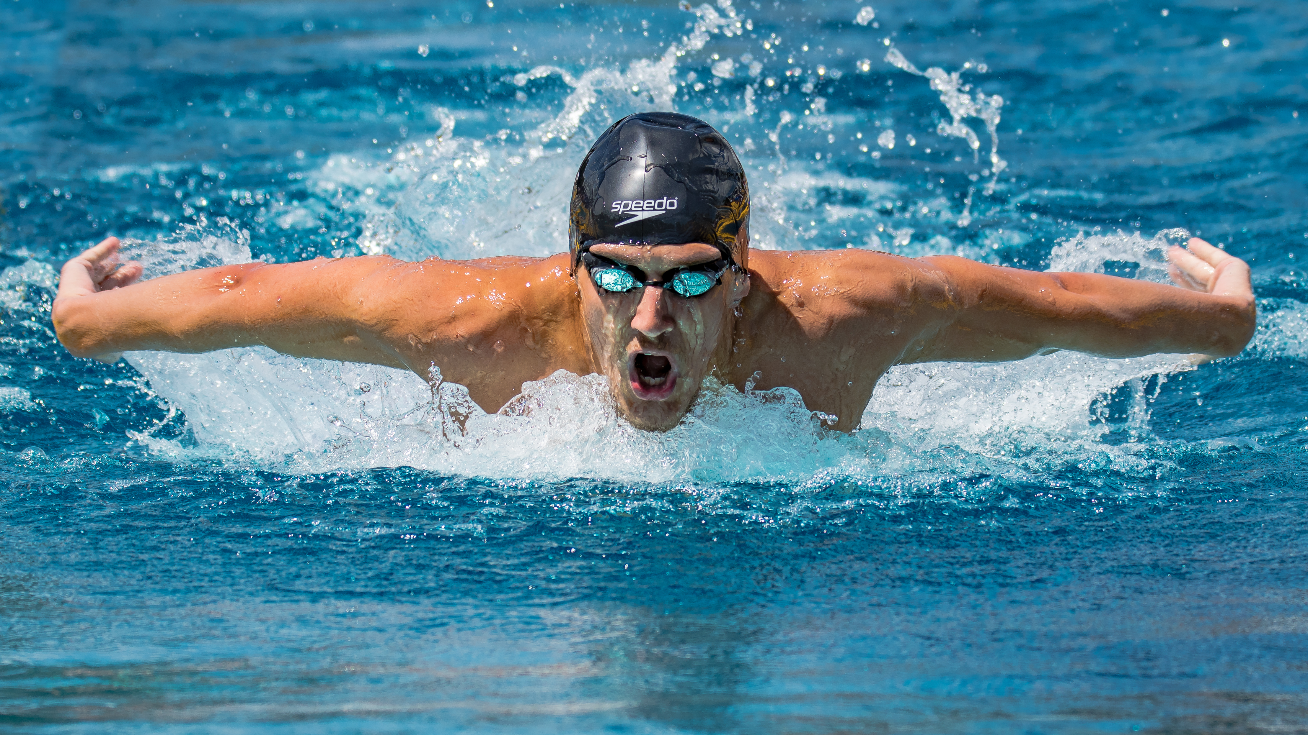 Swimmer in black cap and goggles performs the butterfly stroke, water splashing around outstretched arms.